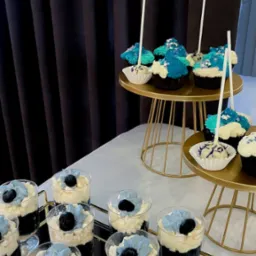 Deserki w szklankach na złotej tacy i babeczki na złotych stojakach, dekorowane w odcieniach błękitu, na tle ciemnej zasłony. Elegancki catering.