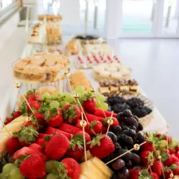 Bogato zdobiony stół z przekąskami: owoce (truskawki, winogrona, arbuz, ananas) na paterze, ciasta i ciasteczka na złotych stojakach. Jasne, naturalne światło.