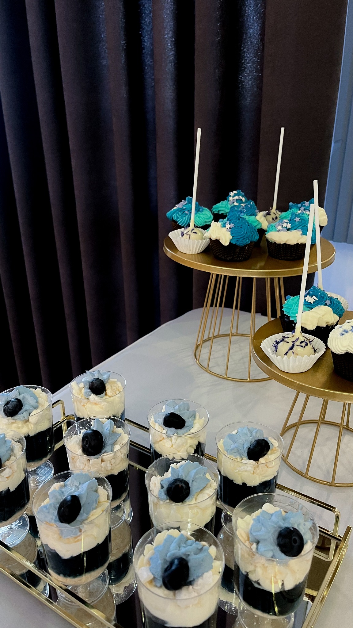 Deserki w szklankach na złotej tacy i babeczki na złotych stojakach, dekorowane w odcieniach błękitu, na tle ciemnej zasłony. Elegancki catering.