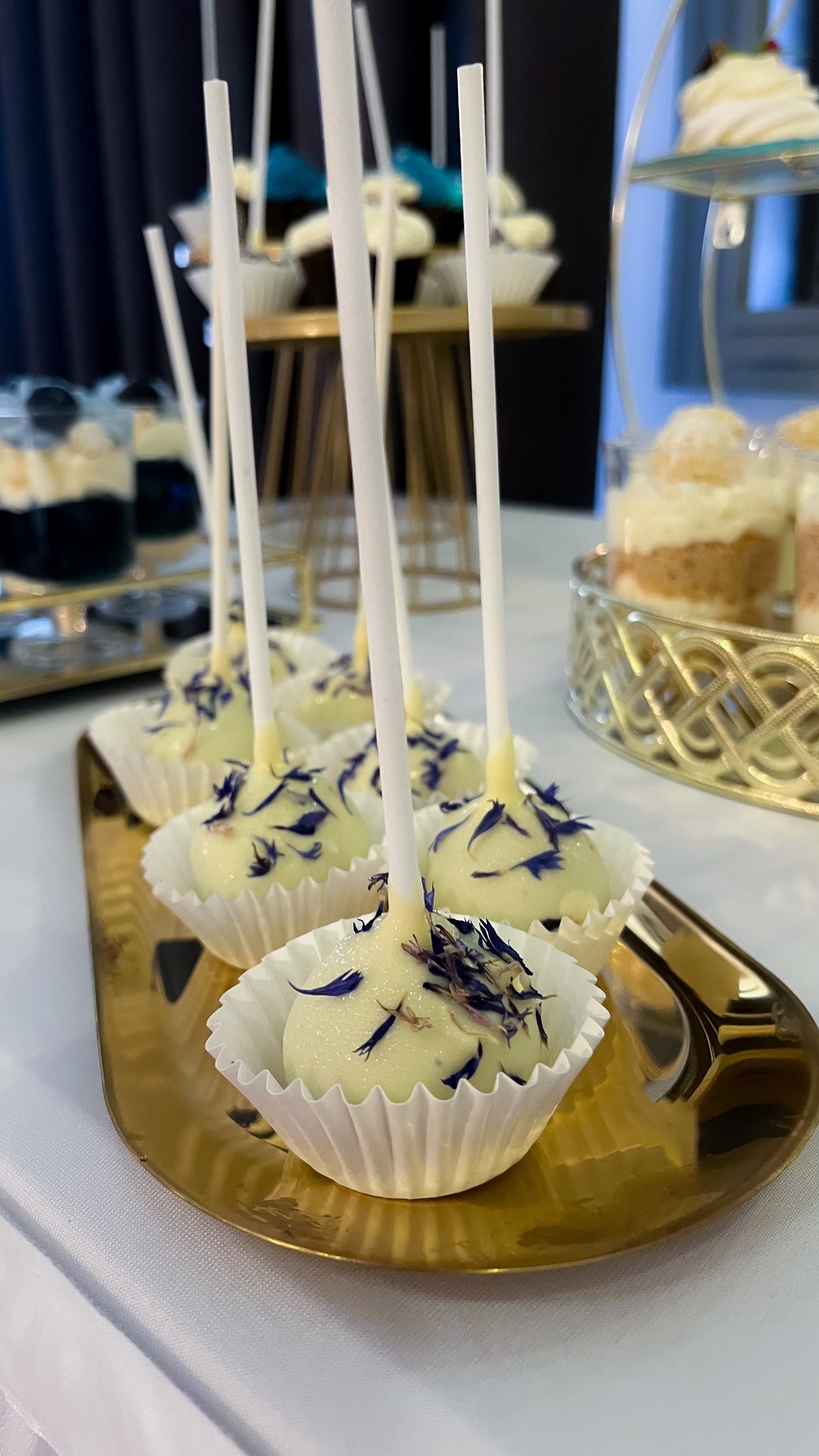 Eleganckie cake pops z białej czekolady i niebieskimi płatkami na złotej tacy, część słodkiego stołu. W tle inne desery.