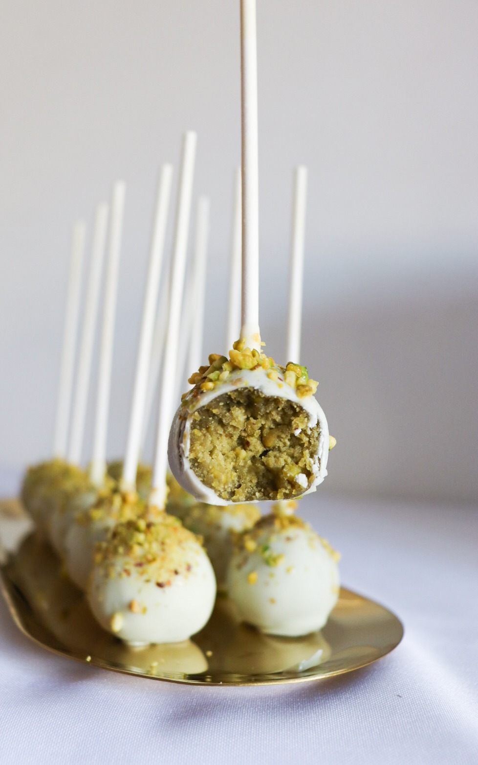 Cake pops z białej czekolady i pistacji na złotym talerzu, jeden przekrojony, ukazujący pistacjowe wnętrze. Delikatne światło podkreśla teksturę.
