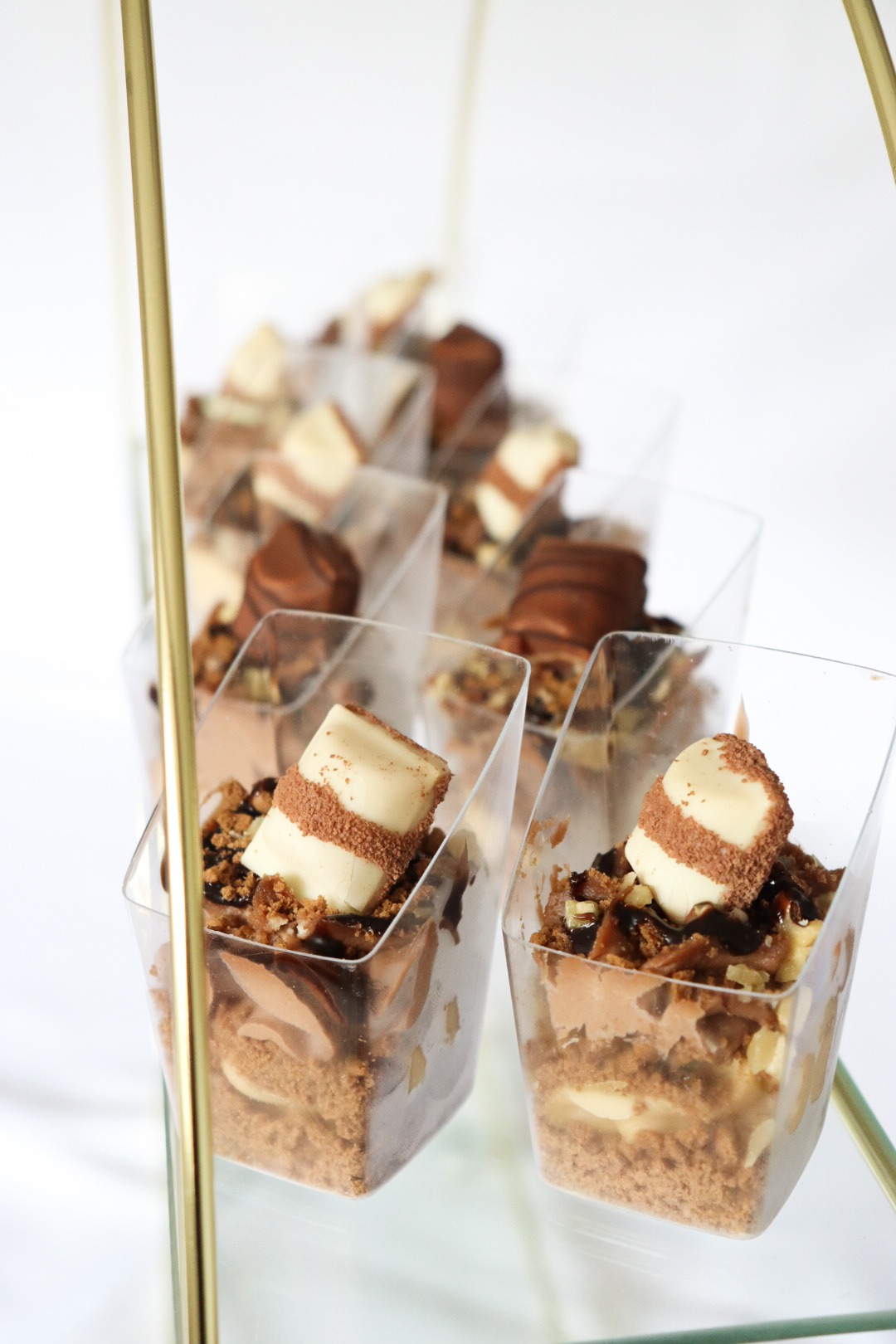 Deserki w przezroczystych, kwadratowych pojemnikach z kawałkami czekolady Kinder na wierzchu, ustawione na szklanym stojaku z elementami w kolorze złota.