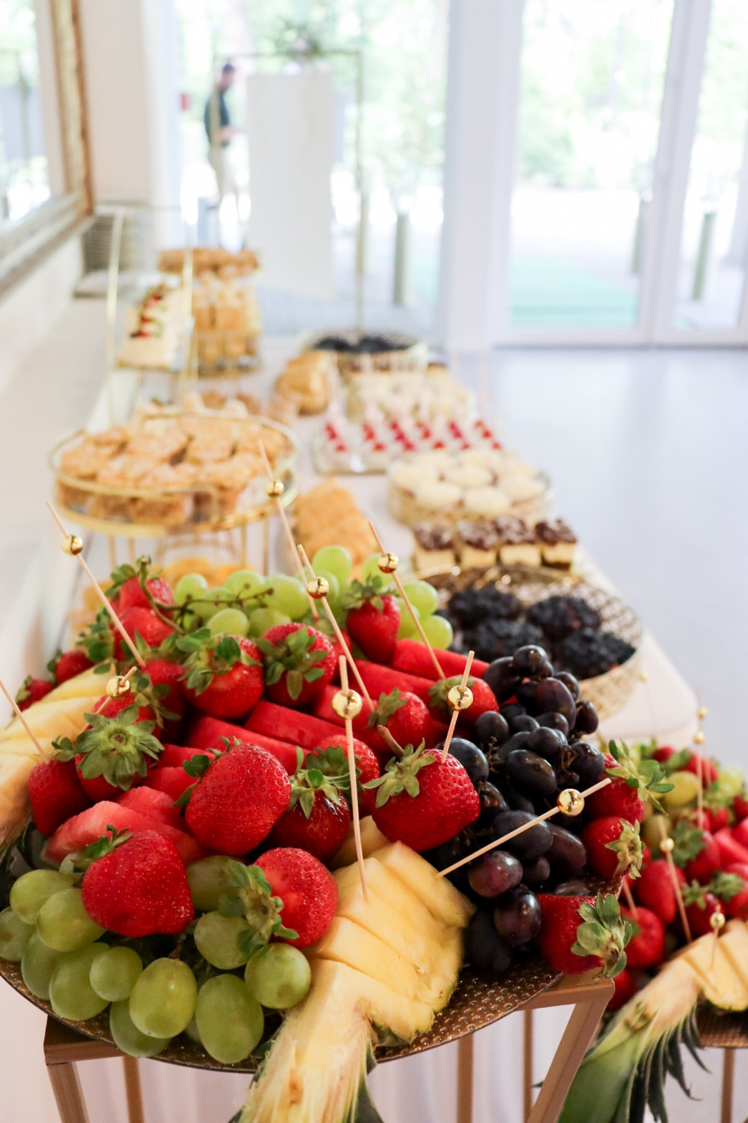 Bogato zdobiony stół z przekąskami: owoce (truskawki, winogrona, arbuz, ananas) na paterze, ciasta i ciasteczka na złotych stojakach. Jasne, naturalne światło.
