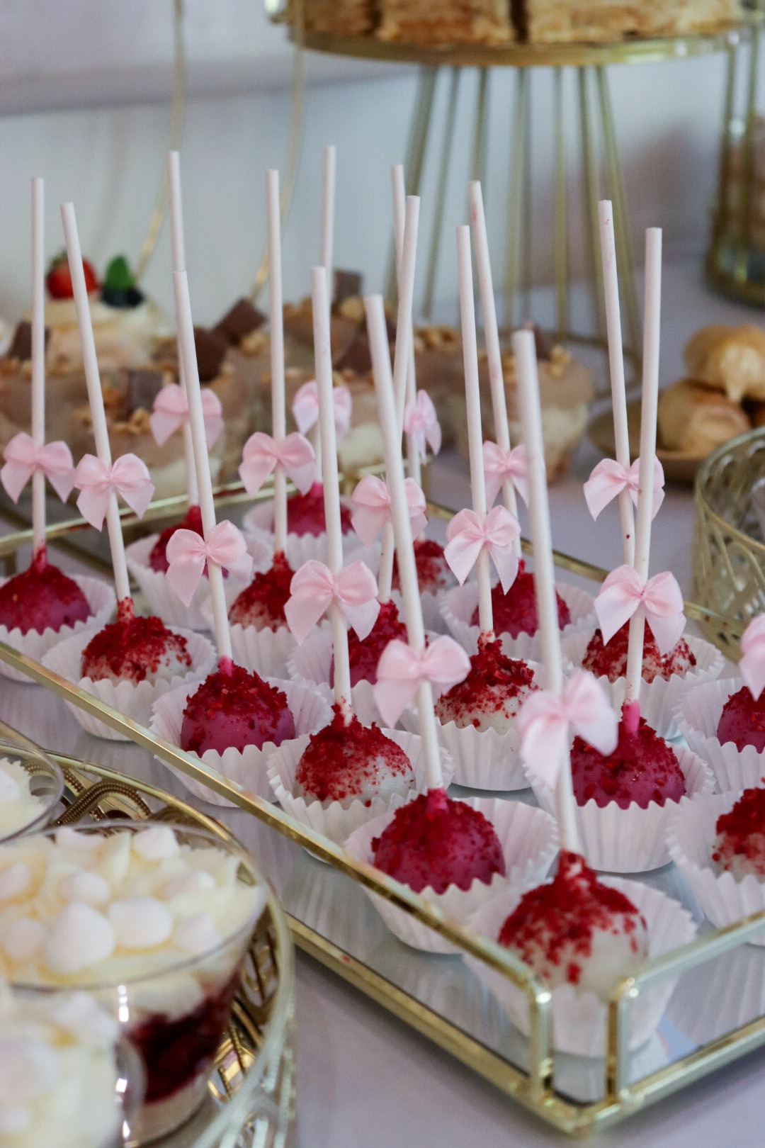 Elegancki srebrny catering: cake pops z różowymi kokardkami i czerwonymi posypkami, deser w szklance z białymi piankami, ciasta w tle na złotym stojaku.