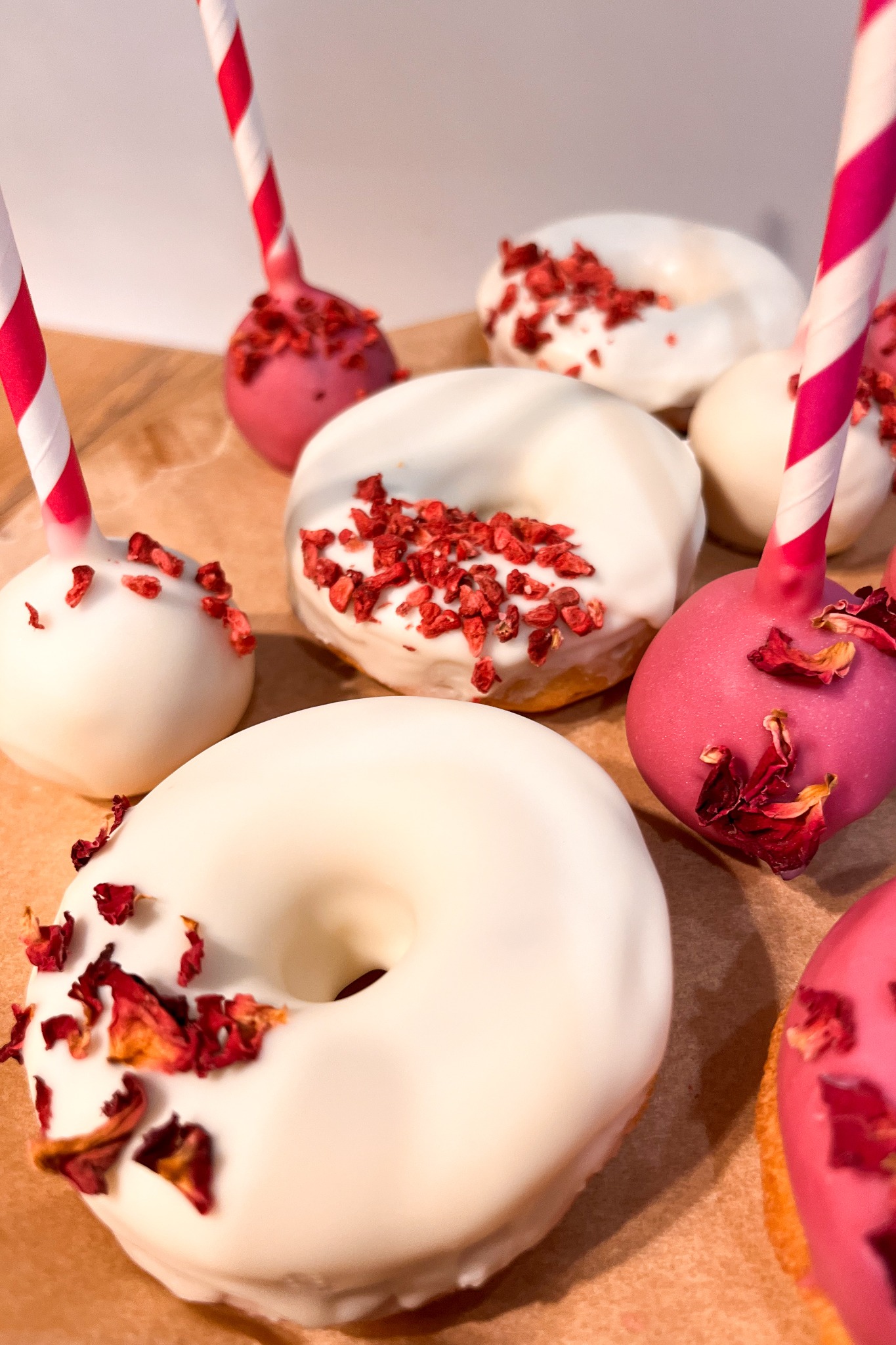 Białe pączki i cake pops w odcieniach różu, udekorowane suszonymi płatkami i czerwonymi posypkami, na papierze pergaminowym.