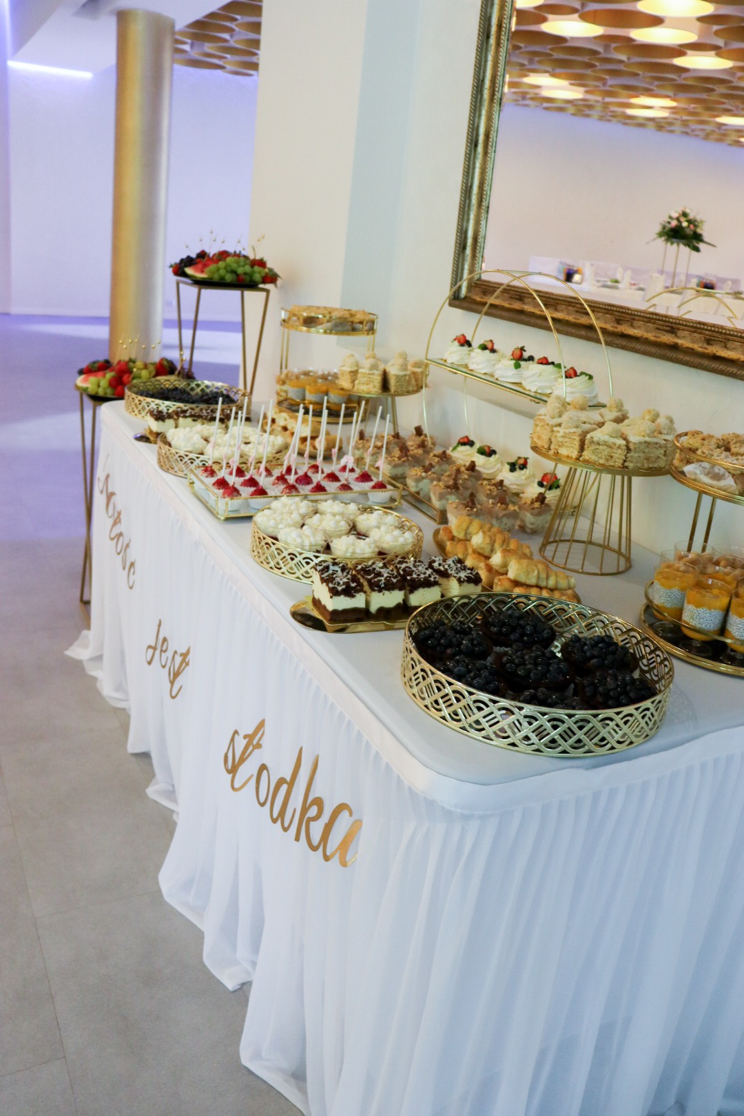 Słodki stół z różnorodnymi deserami na złotych paterach, dekorowany napisem na białym obrusie. Widoczne ciasta, babeczki i owoce.