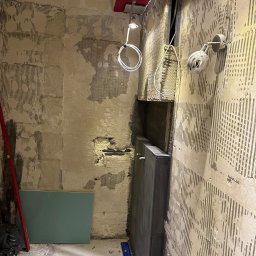 Łukasz Czekała - Wnętrze w trakcie prac wykończeniowych: odsłonięte ściany z resztkami kleju, widoczna instalacja, płyta karton-gips i poziomica. Oświetlenie sufitowe i lampa ścienna.