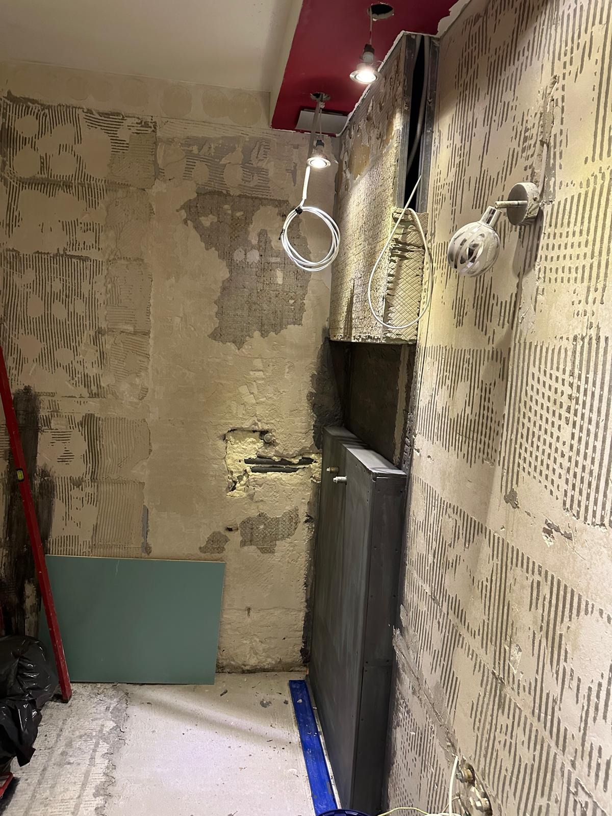 Wnętrze w trakcie prac wykończeniowych: odsłonięte ściany z resztkami kleju, widoczna instalacja, płyta karton-gips i poziomica. Oświetlenie sufitowe i lampa ścienna.