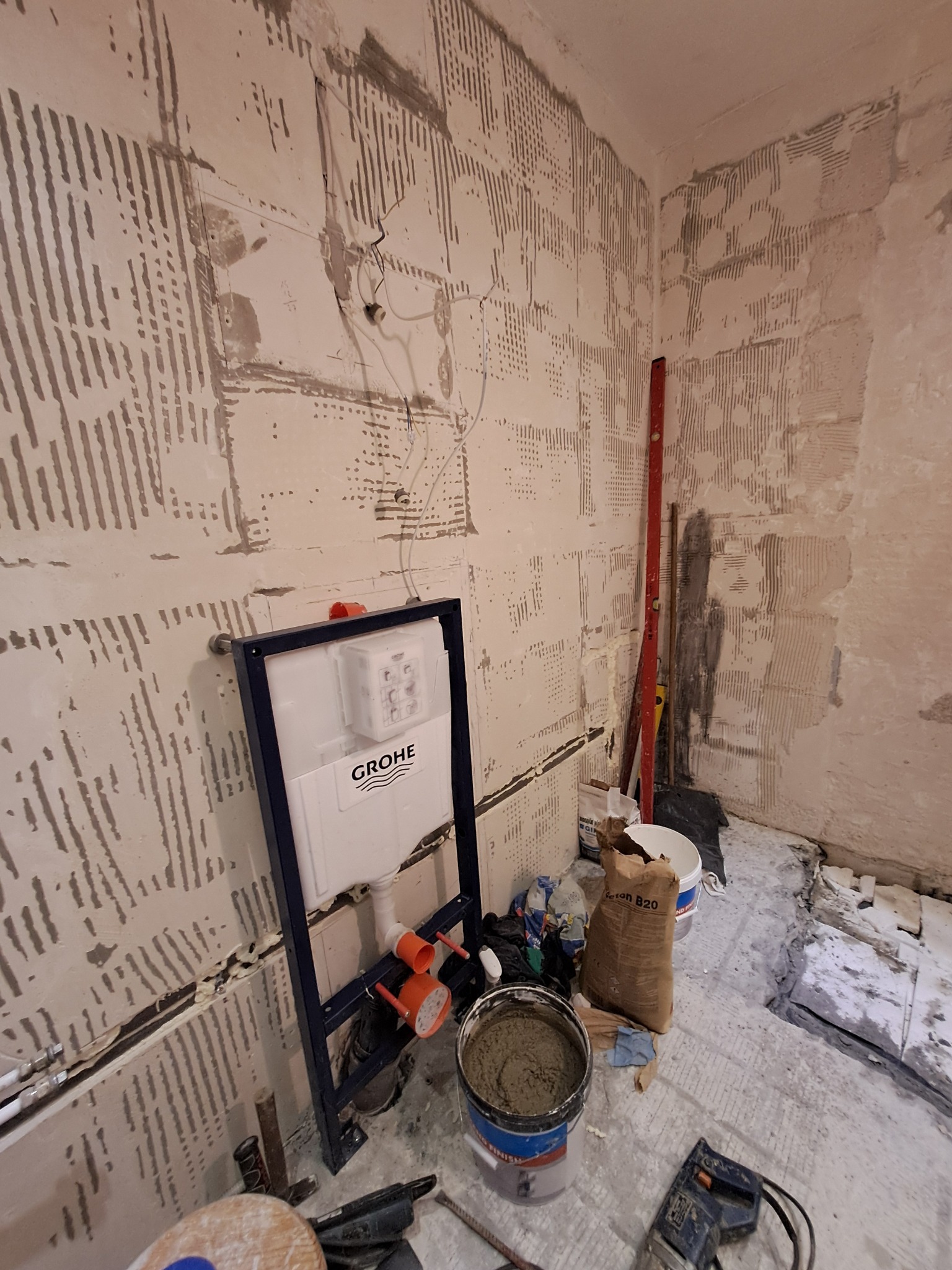 Przygotowanie ściany pod glazurę z zamontowanym stelażem podtynkowym Grohe. Widoczne narzędzia i materiały budowlane. Ściana z charakterystyczną fakturą podłoża.