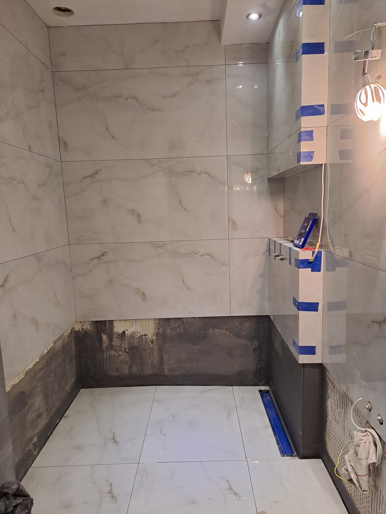 Wnętrze łazienki w trakcie prac glazurniczych. Widoczne białe płytki imitujące marmur na ścianach i podłodze, częściowo położone, z szarym podkładem. W tle półka z niebieską taśmą.