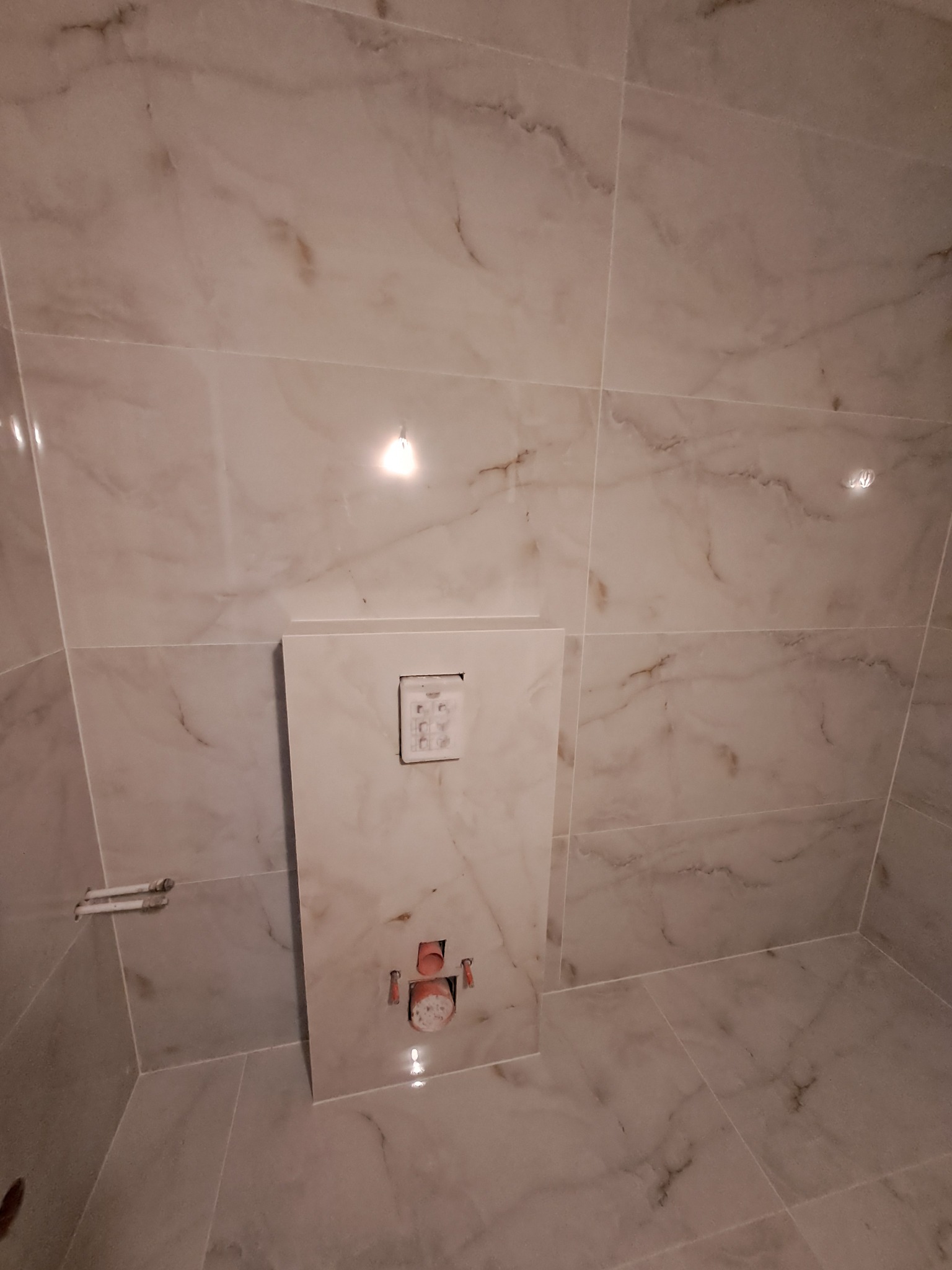 Beżowe płytki imitujące marmur w łazience z zamontowanym stelażem podtynkowym WC i przyciskiem spłukującym. Widoczne przyłącza wodne.