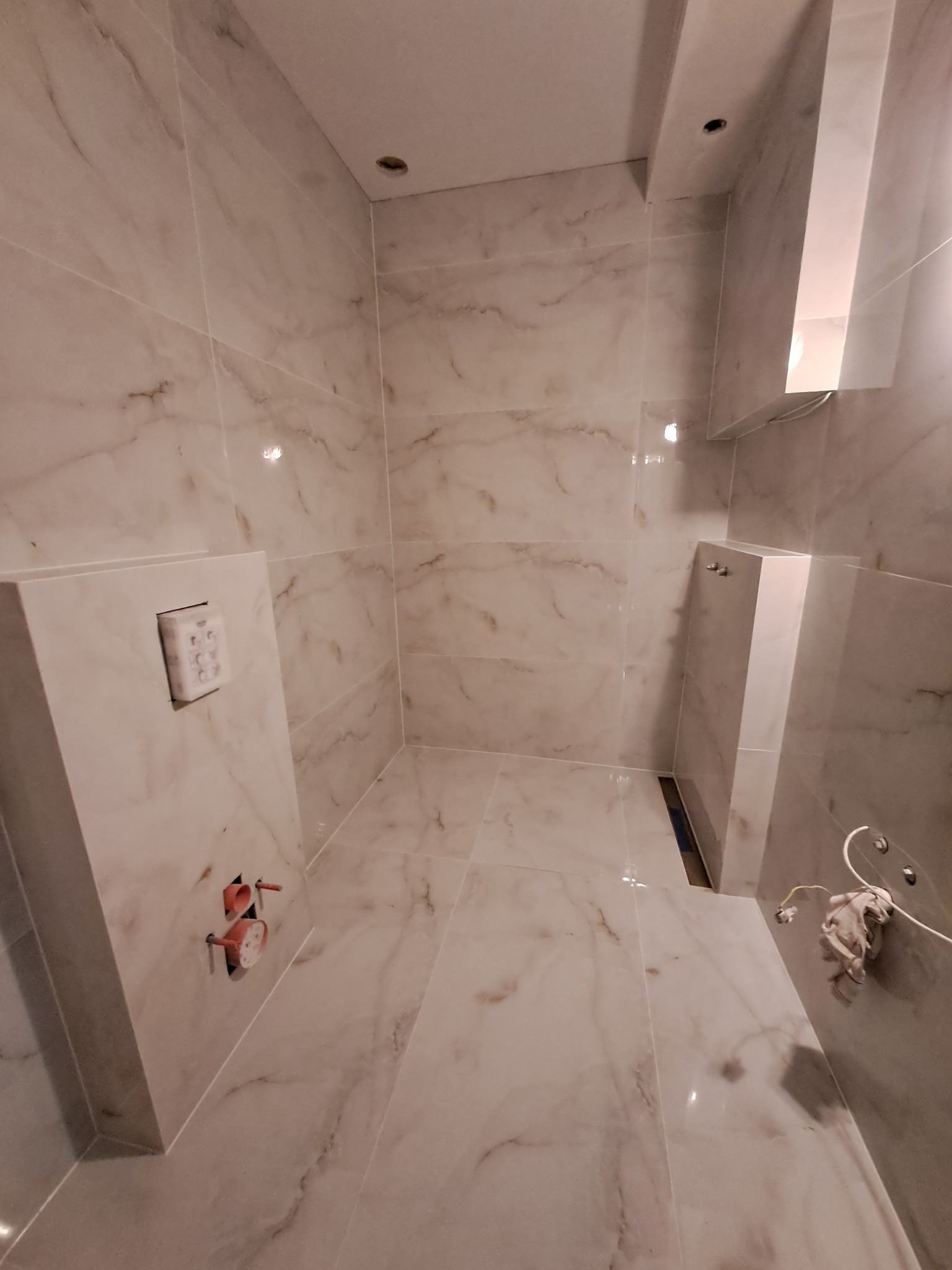 Wykończona łazienka z marmurowymi płytkami, widoczne elementy instalacji wodnej i odpływ liniowy. Minimalistyczny design, jasne oświetlenie.