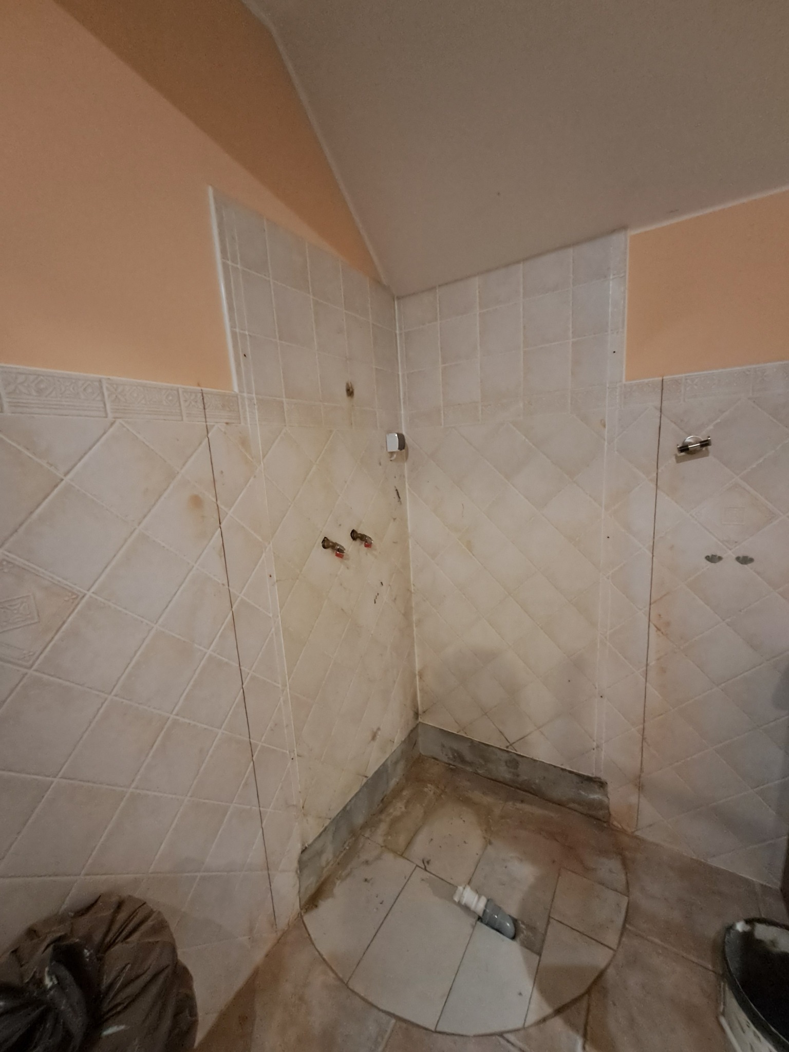 Narożnik łazienki w trakcie prac: częściowo położone płytki w romby, widoczne rury, ślady po kabinie prysznicowej, górna część ściany pomalowana na brzoskwiniowo.