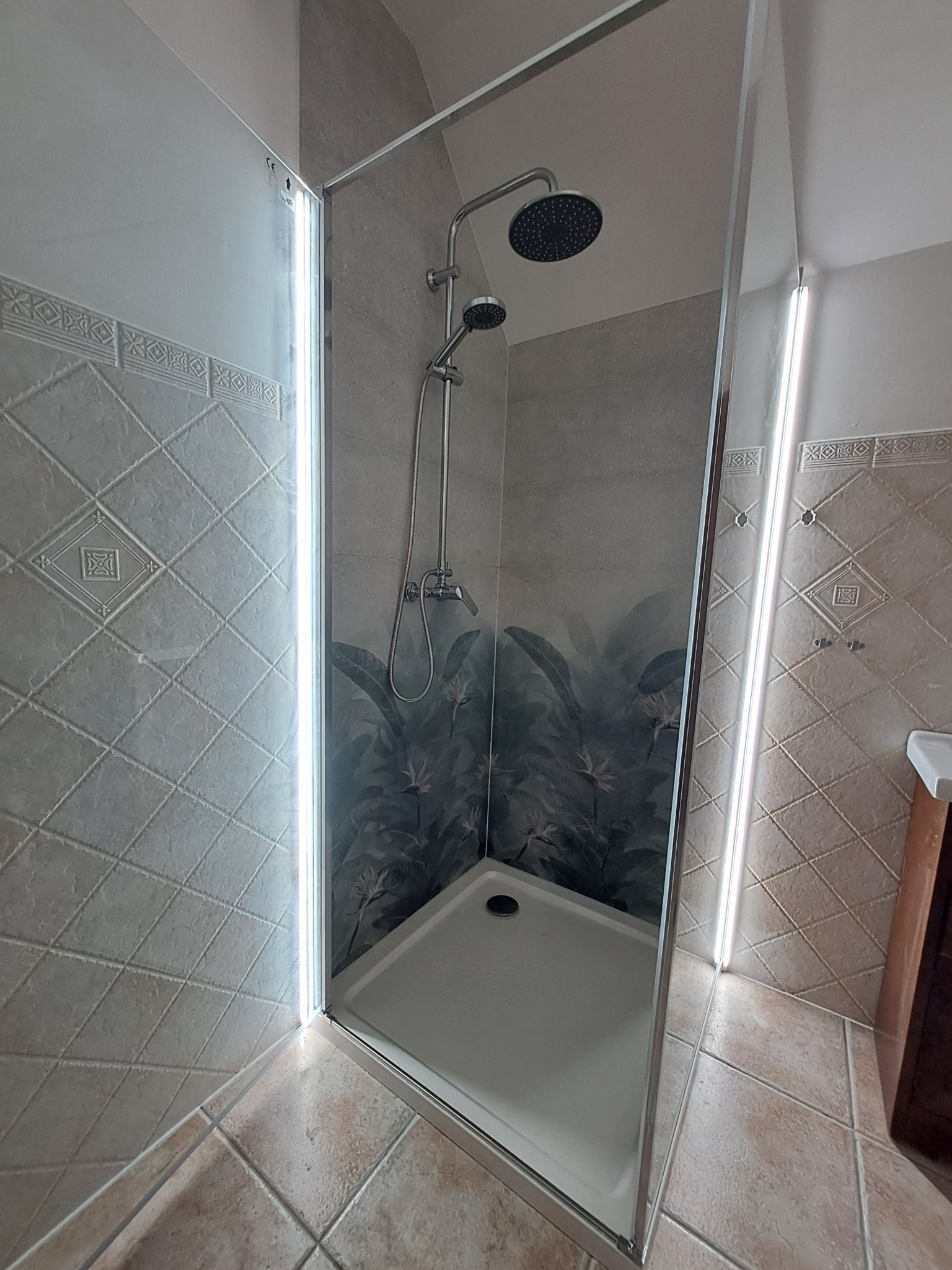 Nowoczesna kabina prysznicowa z szklanymi ścianami, oświetleniem LED i motywem roślinnym na panelu. Widoczne płytki ceramiczne na ścianach i podłodze.