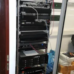 Szafa rack z zainstalowanymi urządzeniami sieciowymi: routery, switche, serwery. Widoczne kable, panele krosownicze i zasilacze awaryjne APC. Uporządkowana instalacja.