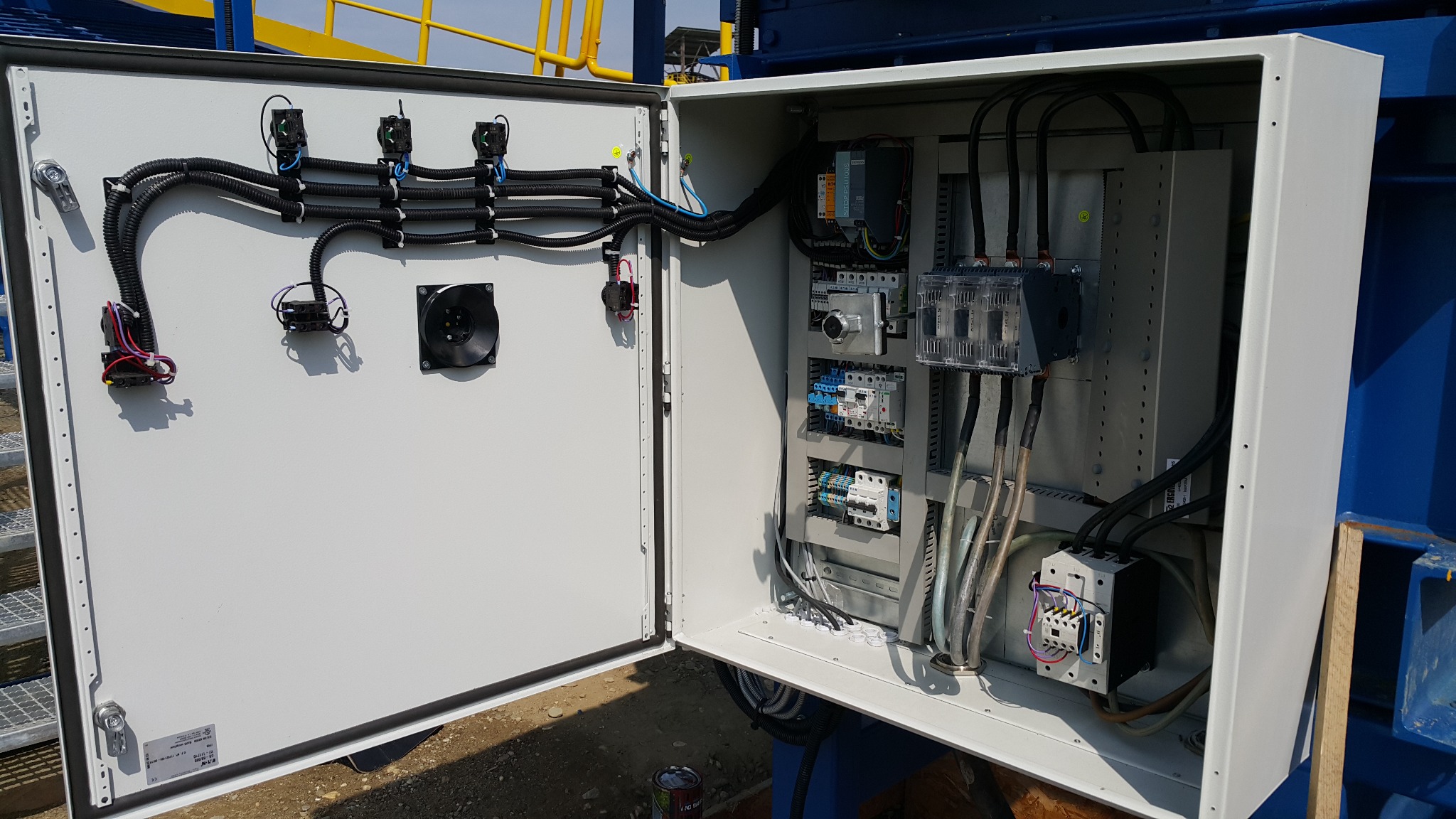 Otwarta szafa sterownicza z widocznymi elementami elektrycznymi, przewodami i wentylatorem. Szyna DIN z zabezpieczeniami i przekaźnikami wewnątrz obudowy.