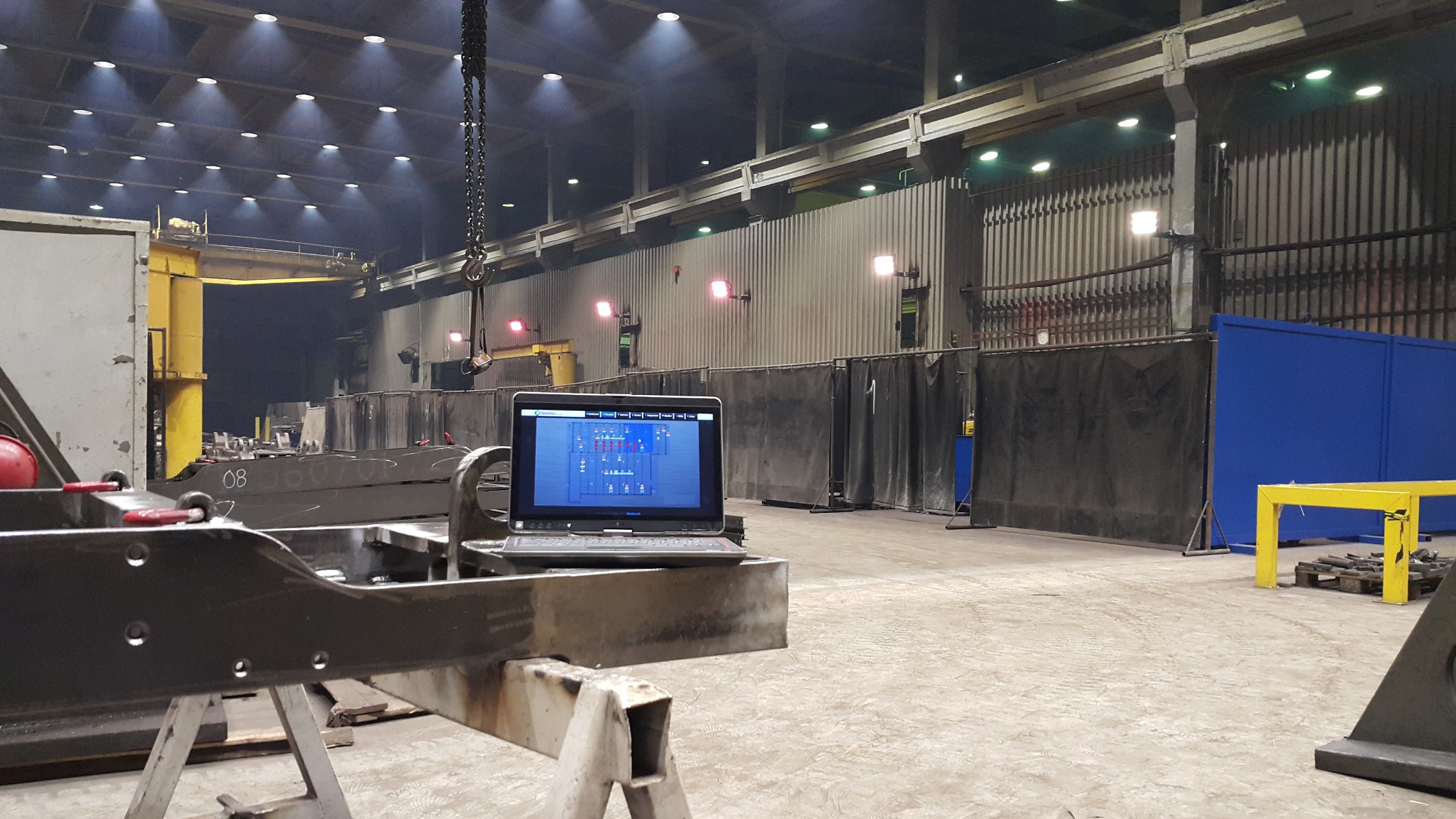 Laptop z oprogramowaniem sterującym na tle hali produkcyjnej z suwnicą i elementami konstrukcyjnymi. Widoczne oświetlenie przemysłowe i stanowiska pracy.