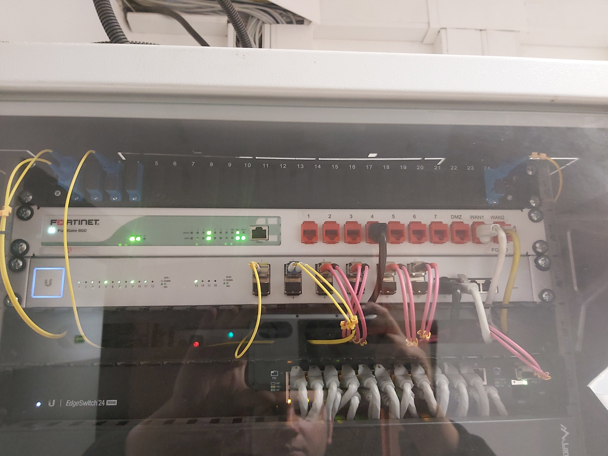 Szafa rack z zainstalowanymi urządzeniami sieciowymi: Fortinet, EdgeSwitch. Widoczne kable Ethernet w różnych kolorach. Porty ponumerowane. Dioda sygnalizacyjna świeci.