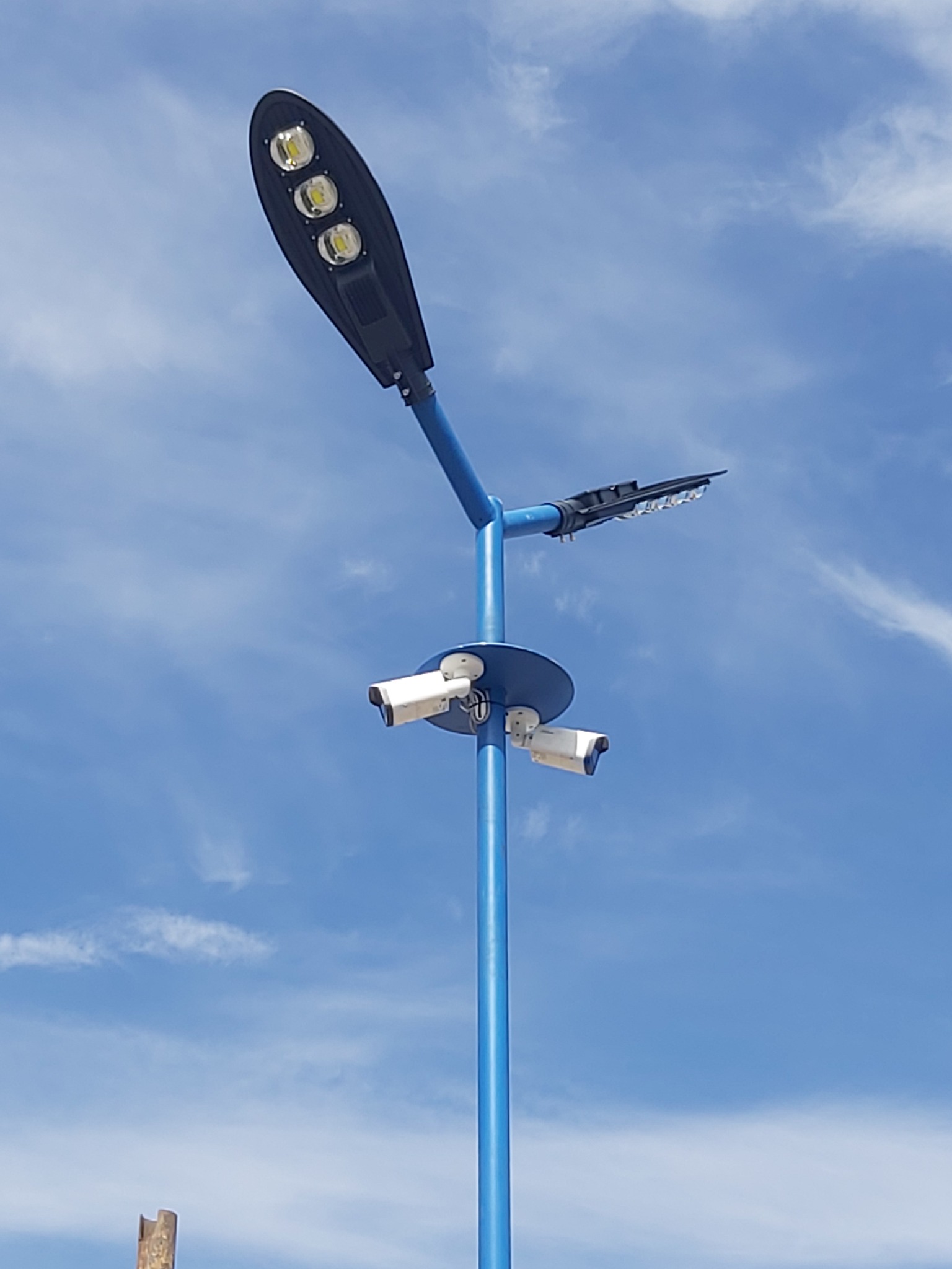 Niebieski słup oświetleniowy z trzema kamerami monitoringu i panelem LED, na tle błękitnego nieba z chmurami.