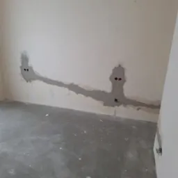 Wnętrze w trakcie prac przygotowawczych: ściana z wyfrezowanymi bruzdami pod instalacje elektryczne, wypełnionymi szarą masą. Surowa podłoga, wisząca żar&oacute;wka.