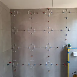 Ułożone płytki w łazience, szare, z systemem poziomowania. Widoczne otwory na instalacje, puszka elektryczna i fragment okna. Prace glazurnicze w toku.