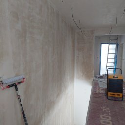 artis - Ściana po szpachlowaniu, z widocznym wałkiem malarskim opartym o ścianę. W tle drabina i nagrzewnica Master. Widoczne kable wystające ze ściany.