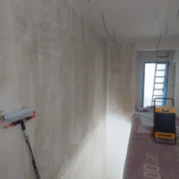 Ściana po szpachlowaniu, z widocznym wałkiem malarskim opartym o ścianę. W tle drabina i nagrzewnica Master. Widoczne kable wystające ze ściany.