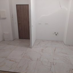 artis - Wykończone wnętrze z beżową podłogą imitującą marmur, białe ściany z instalacjami i brązowe drzwi z metalowymi elementami w nowym mieszkaniu.
