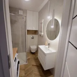 Nowoczesna łazienka z kabiną prysznicową, umywalką z szafką i drewnopodobną podłogą ułożoną w jodełkę. Oświetlenie punktowe i okrągłe lustro z podświetleniem LED.