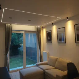 Nowoczesny salon z narożnikiem i oświetleniem LED na suficie, widok na ogród przez przeszklone drzwi balkonowe. Minimalistyczny design z czarnymi lampami punktowymi.