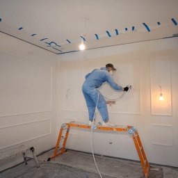 artis - Pracownik w niebieskim kombinezonie i masce natryskowo pokrywa białą ścianę z ozdobnymi sztukateriami, stojąc na pomarańczowym rusztowaniu w remontowanym wnętrzu.