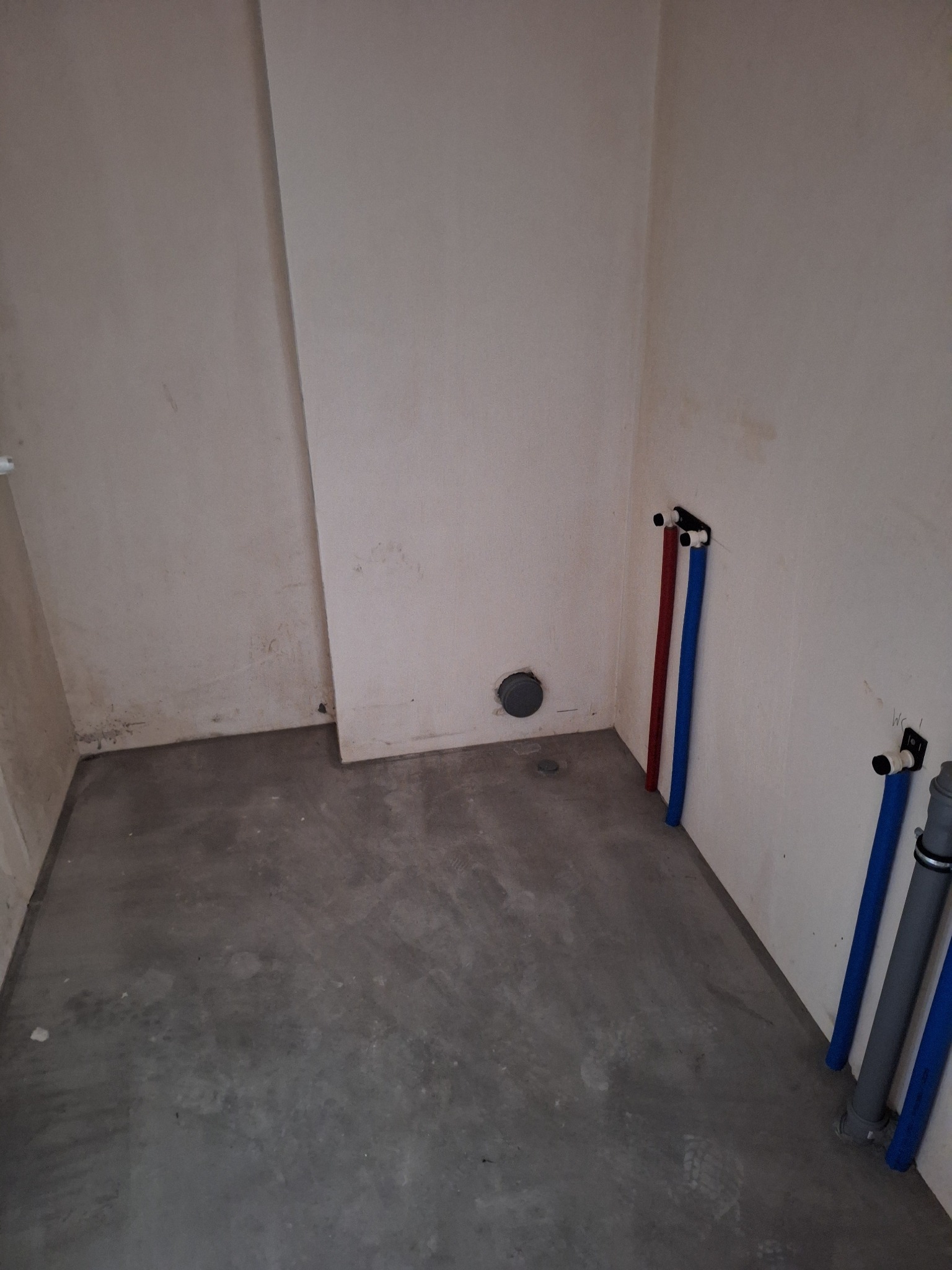 Surowy stan łazienki w trakcie remontu: widoczne rury instalacji wodnej w kolorze czerwonym i niebieskim, szara wylewka i ściany przygotowane do dalszych prac.
