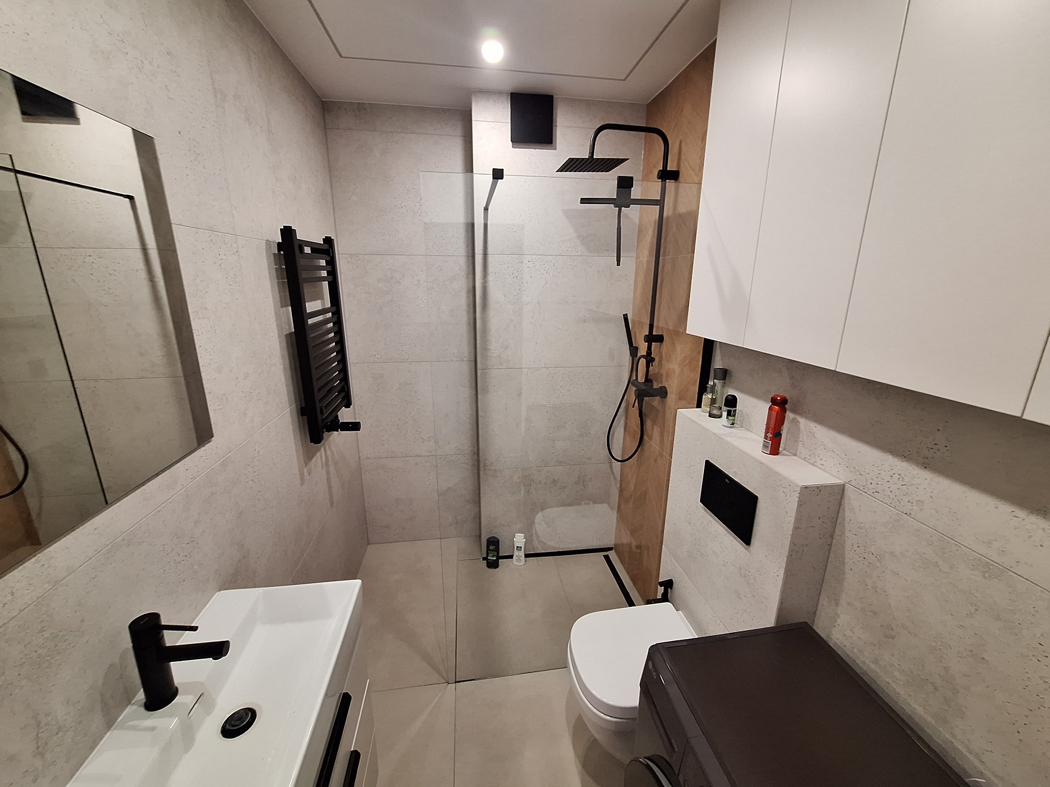 Nowoczesna łazienka z kabiną typu walk-in, czarną armaturą i grzejnikiem drabinkowym. Umywalka nablatowa i szafki w bieli. Stonowana kolorystyka płytek.