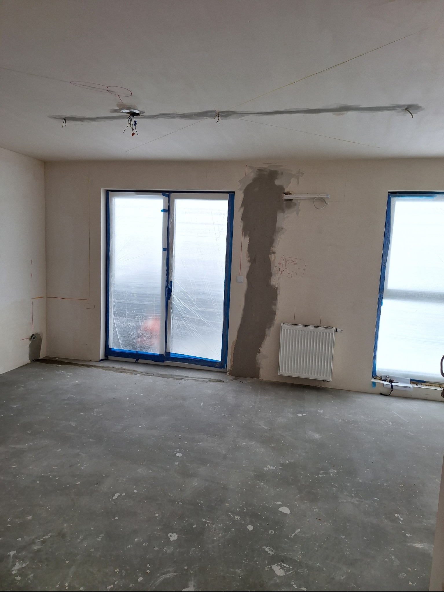 Wnętrze w trakcie prac: ściany po szpachlowaniu, okna zabezpieczone folią, widoczne przewody elektryczne na suficie i grzejnik. Przygotowanie do dalszych prac wykończeniowych.