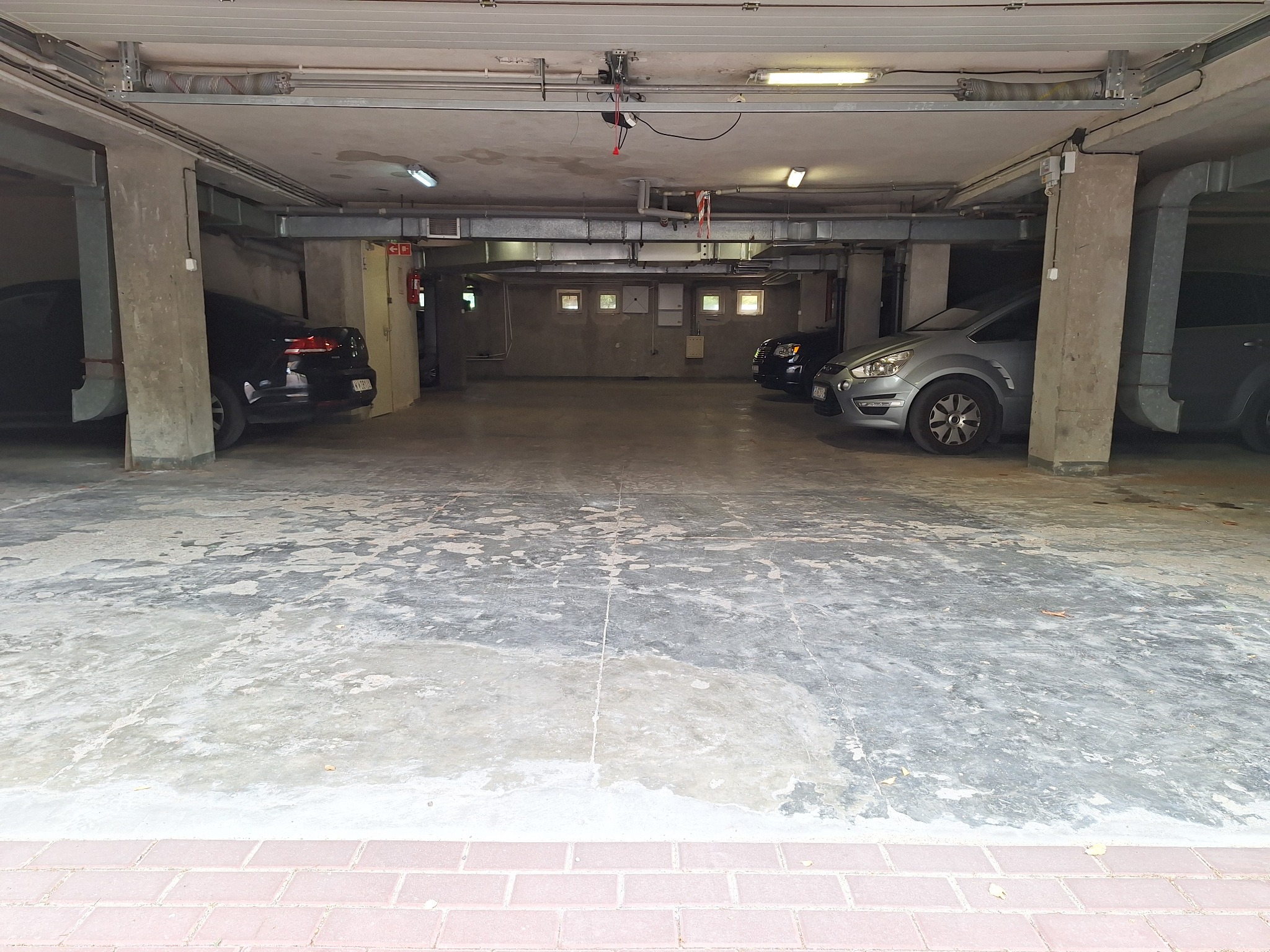 Wnętrze garażu podziemnego z betonową posadzką i widocznymi elementami instalacji. Ujęcie z poziomu posadzki, w tle zaparkowane samochody.