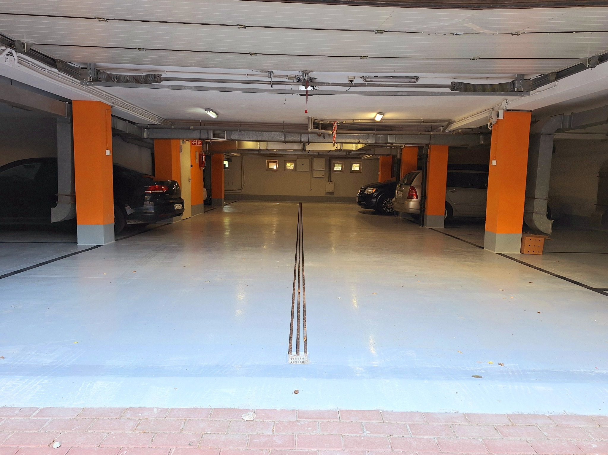 Wnętrze garażu podziemnego z pomalowaną na szaro posadzką i pomarańczowymi kolumnami. Widoczne samochody po bokach i elementy instalacji pod sufitem.