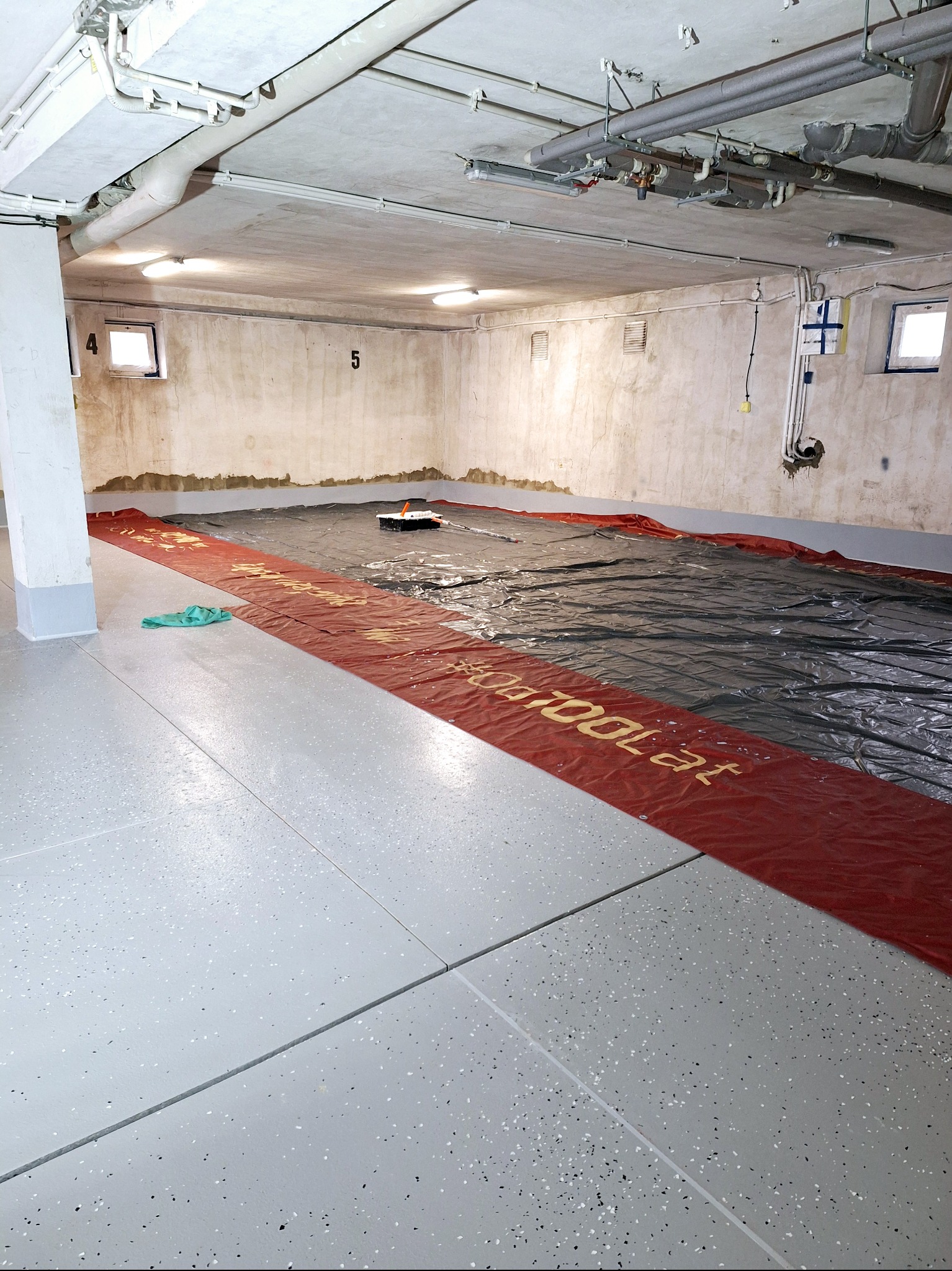 Biało-szara posadzka w piwnicy, zabezpieczona folią i taśmą malarską, przygotowana do dalszych prac wykończeniowych. Widoczne rury pod sufitem i surowe ściany.