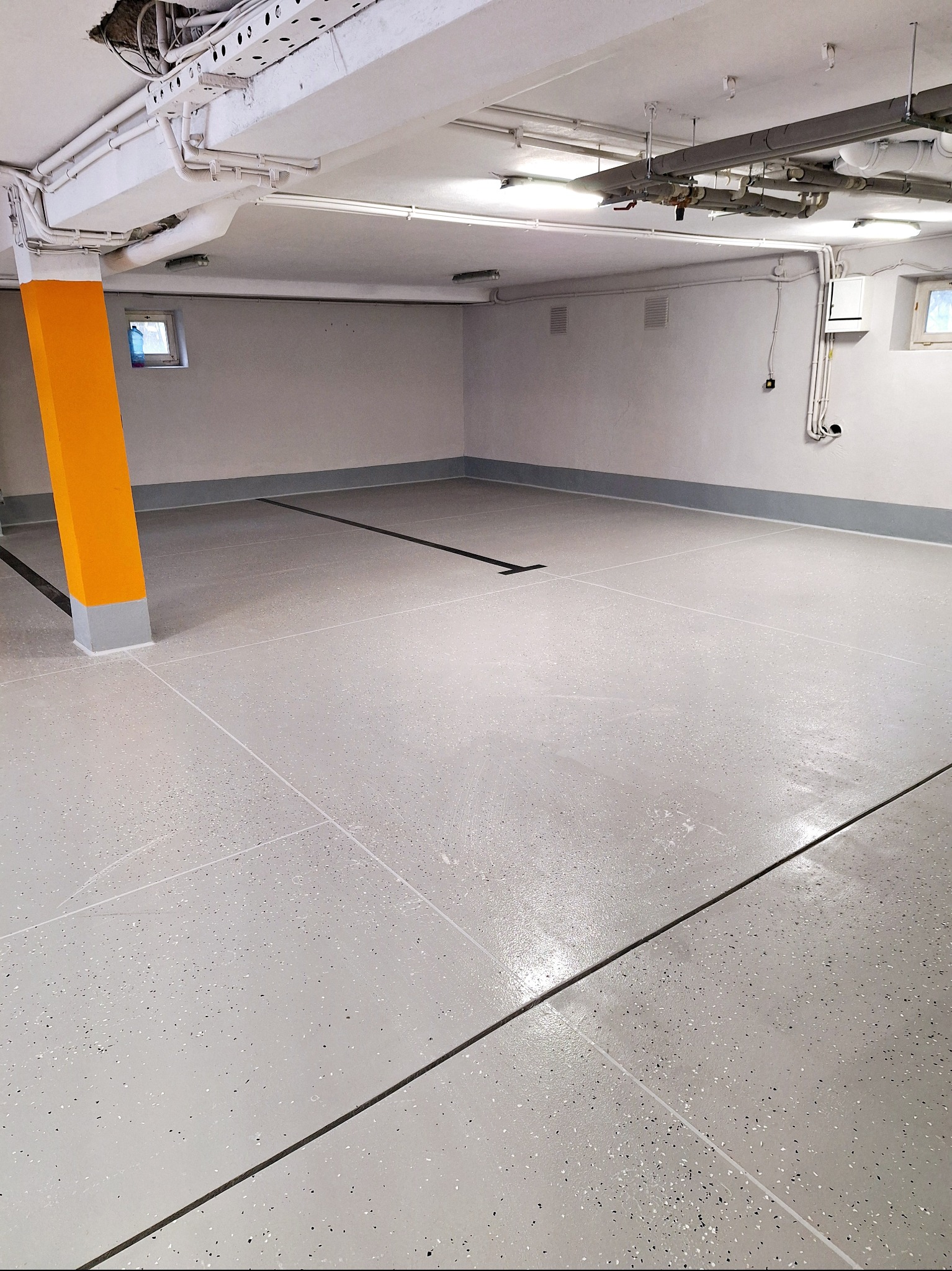 Jasnoszara posadzka żywiczna z ciemnymi akcentami w przestronnym, odnowionym garażu lub pomieszczeniu gospodarczym z pomalowanym na żółto słupem.