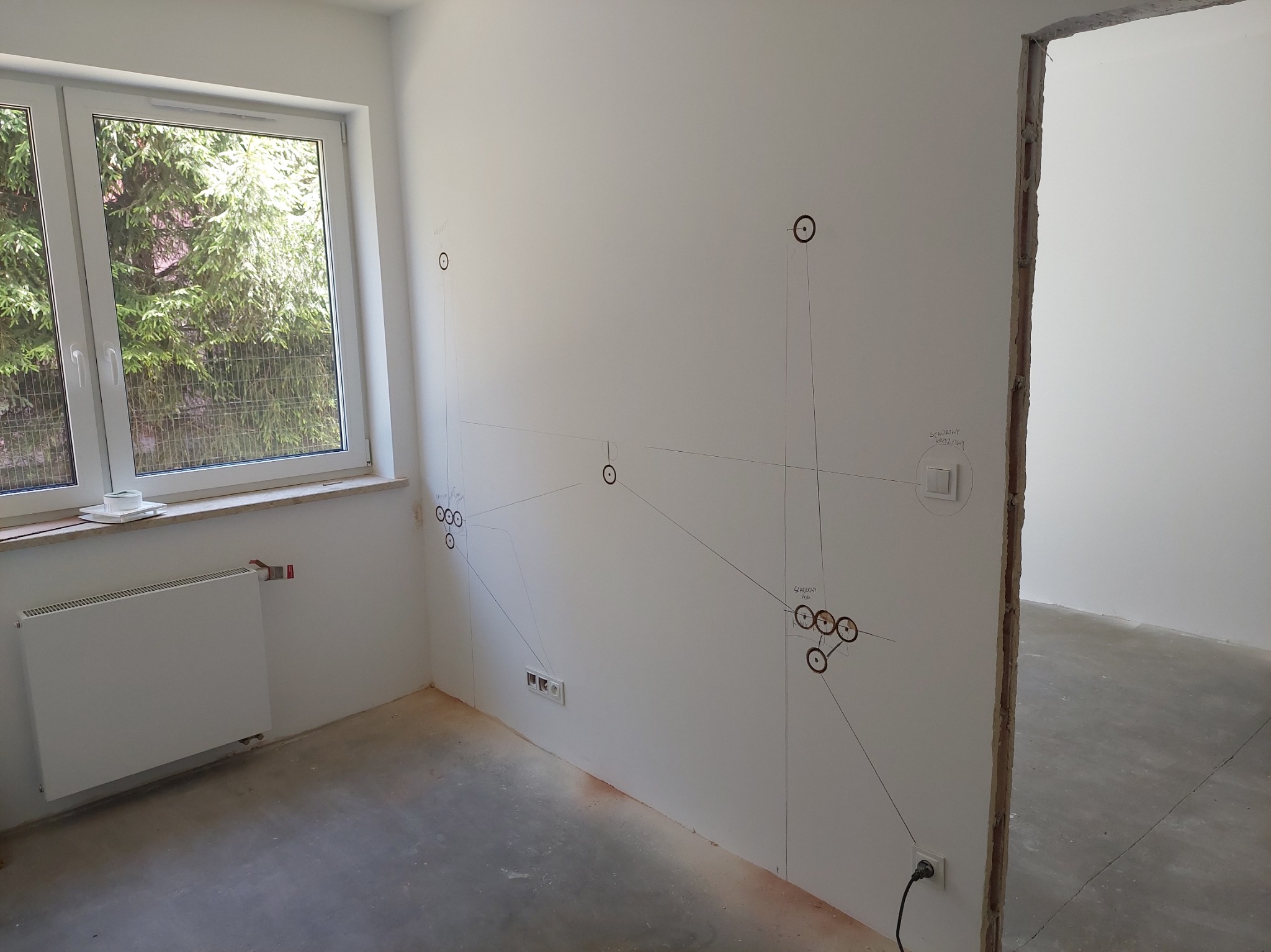 Białe wnętrze w trakcie prac wykończeniowych. Na ścianie widoczne oznaczenia instalacji elektrycznej. Okno z widokiem na zieleń. Stan surowy podłogi.