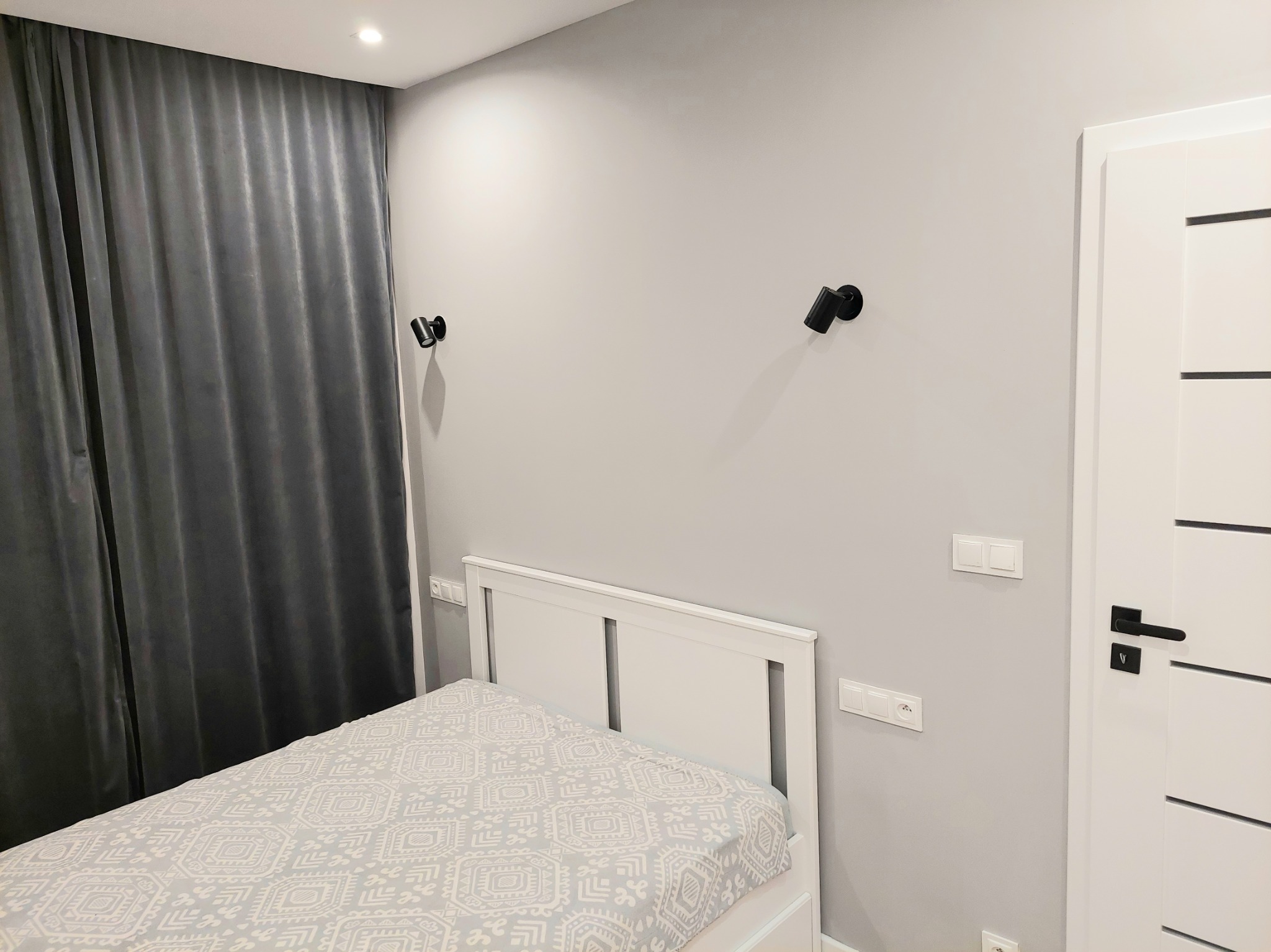 Jasnoszara sypialnia z łóżkiem, czarnymi lampkami ściennymi i białą, nowoczesną stolarką drzwiową. Szare zasłony w tle.