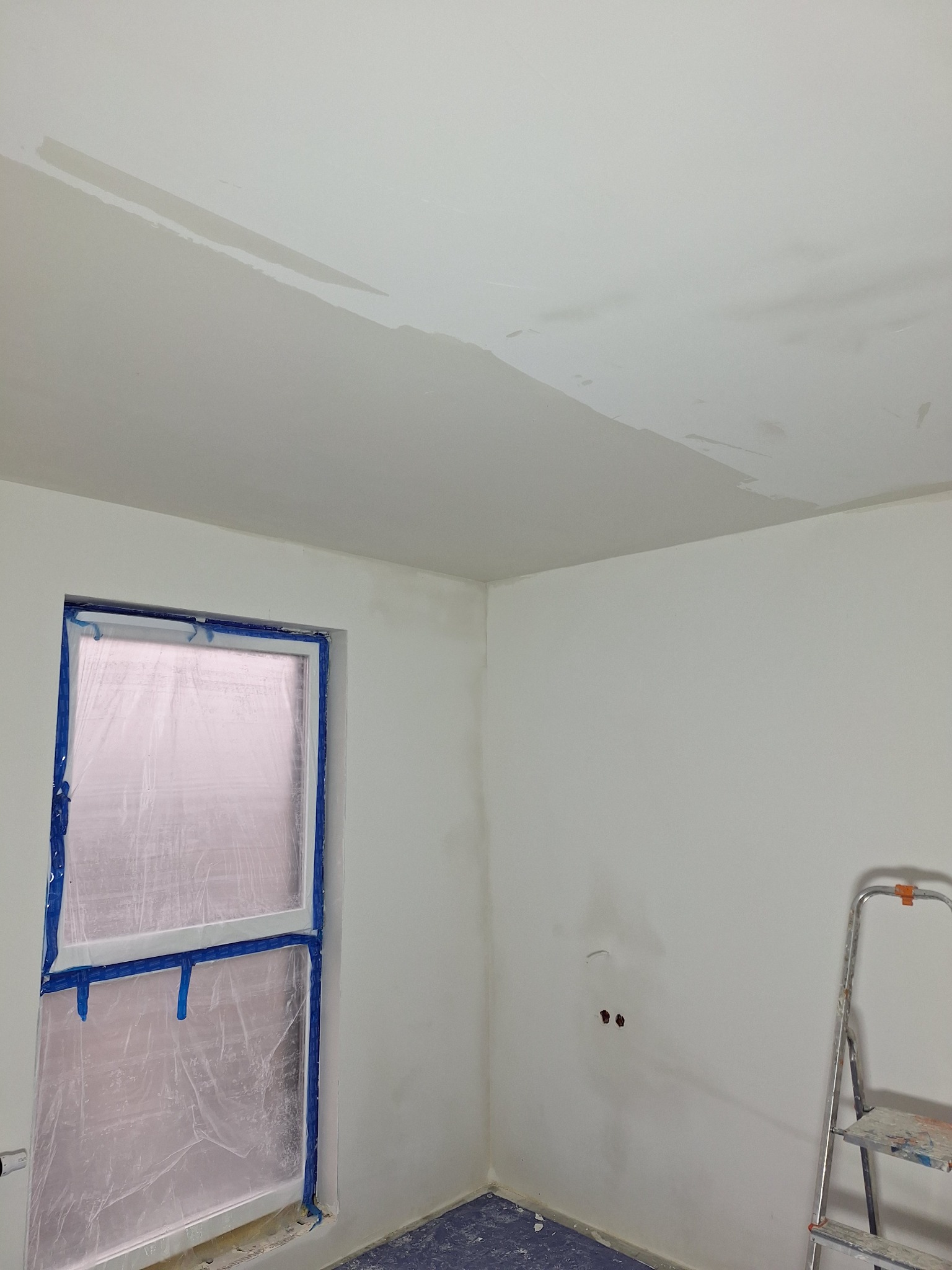 Pokój w trakcie prac przygotowawczych: okno zabezpieczone folią i taśmą, częściowo wyszpachlowany sufit, drabina malarska w rogu.