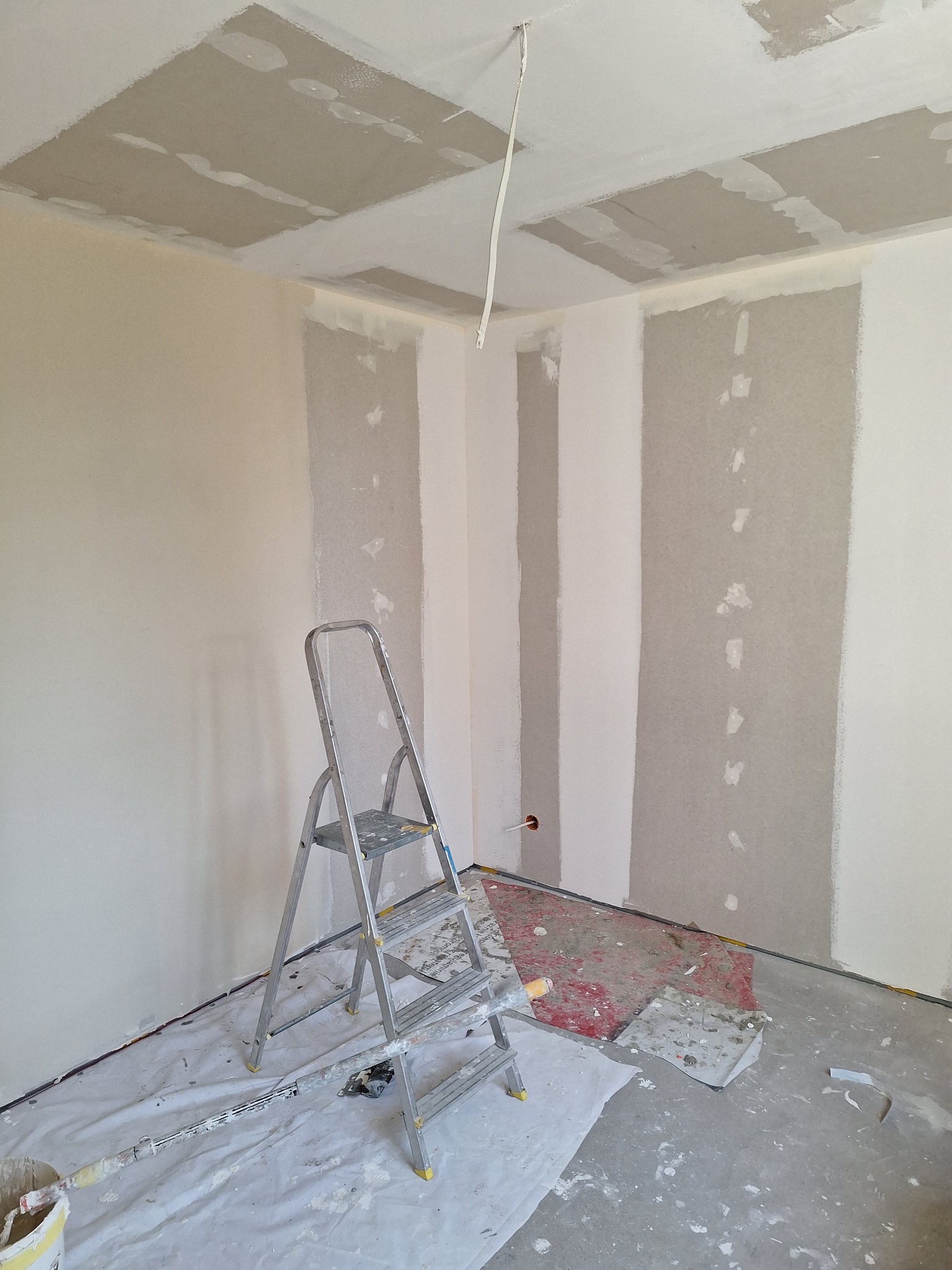 Pokój w trakcie szpachlowania: częściowo wyszpachlowane ściany, drabina aluminiowa, zabezpieczona podłoga. Widoczne niedoskonałości i surowość wykończenia.