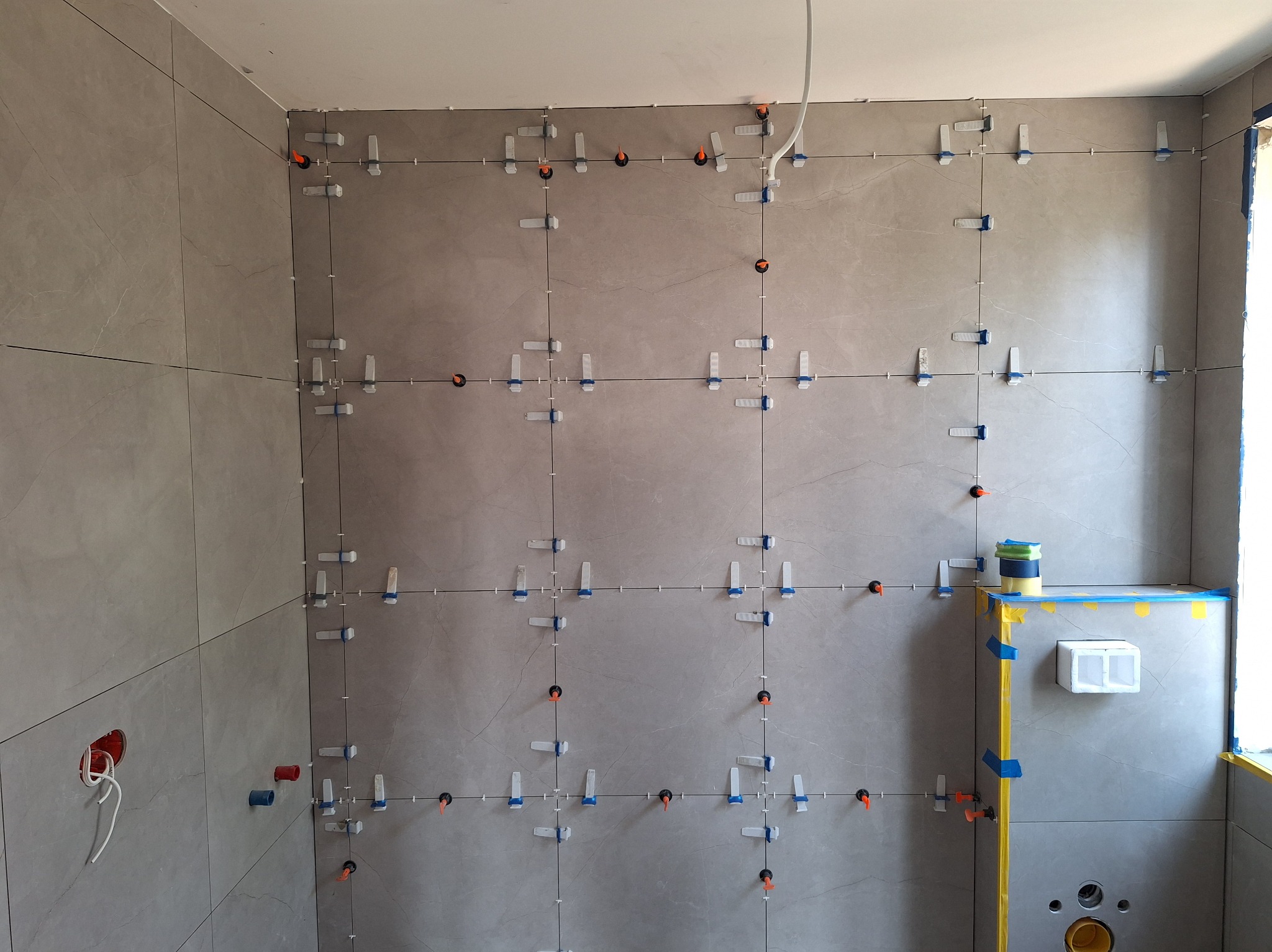 Ułożone płytki w łazience, szare, z systemem poziomowania. Widoczne otwory na instalacje, puszka elektryczna i fragment okna. Prace glazurnicze w toku.