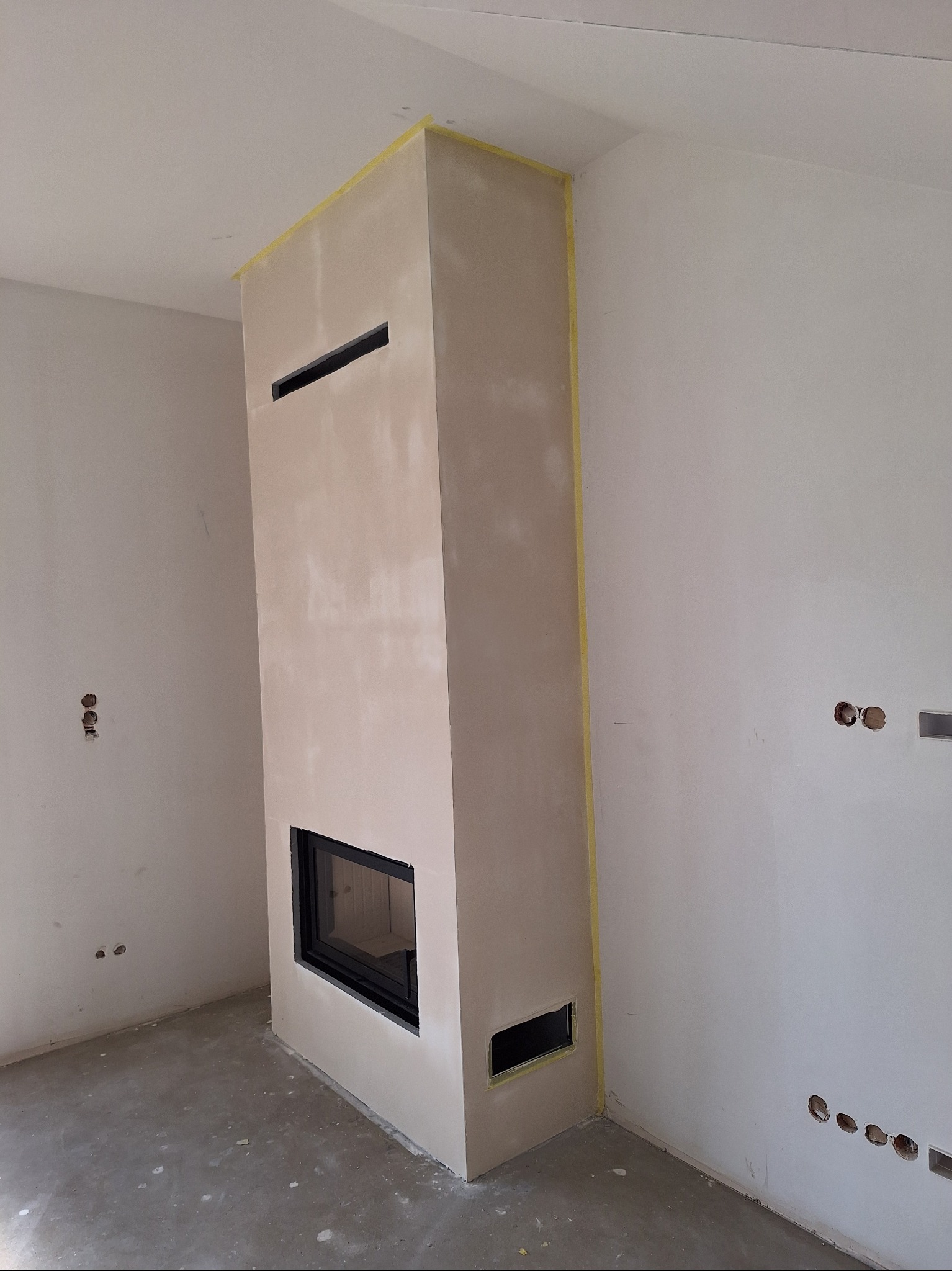 Wysoki kominek obudowany płytami, zabezpieczony taśmą malarską, w surowym stanie. Widoczne otwory wentylacyjne i wkład kominkowy. Podłoga surowa, ściany przygotowane do malowania.