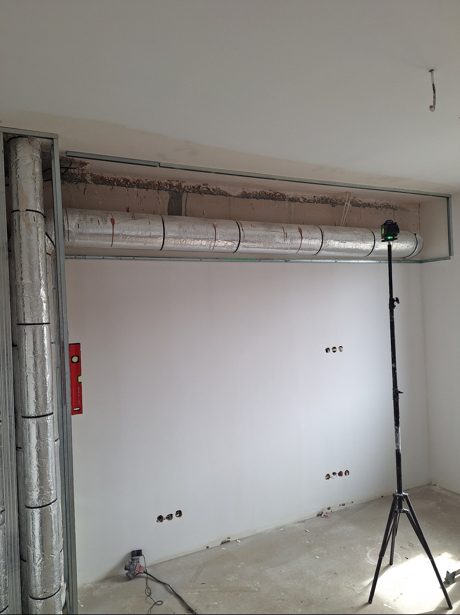 Wnętrze w trakcie prac wykończeniowych: widoczna izolacja rur pod sufitem w metalowej konstrukcji, poziomica, laser krzyżowy na statywie i ślady po instalacji elektrycznej.