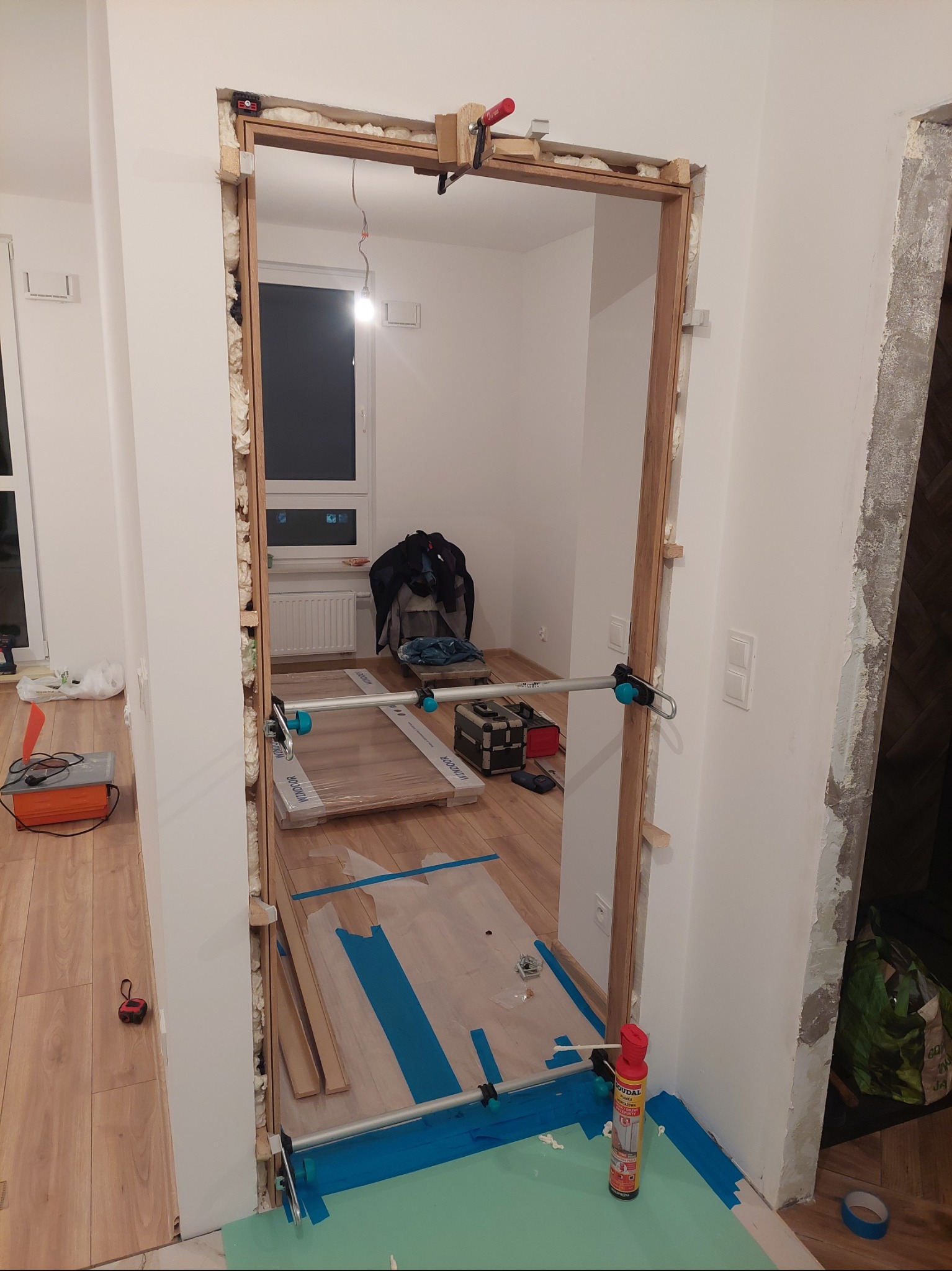 Montaż ościeżnicy drzwiowej z użyciem pianki montażowej i rozpórek. Widoczne przygotowane panele podłogowe i akcesoria montażowe.