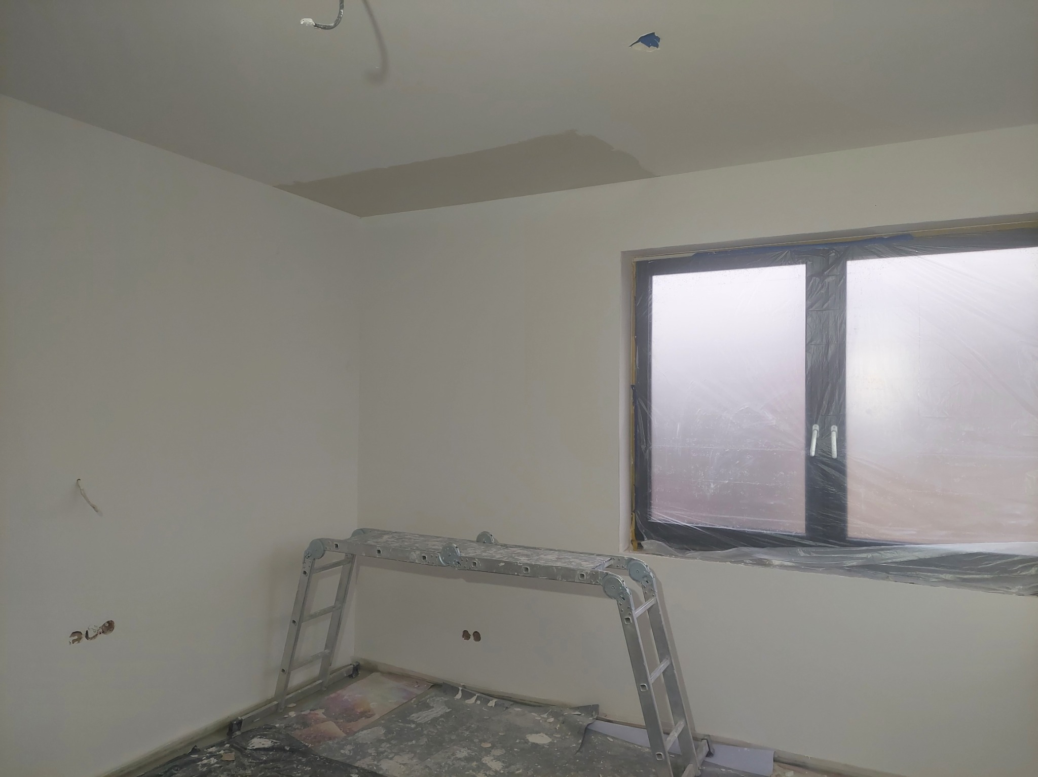 Pokój w trakcie prac wykończeniowych: ściany pomalowane na biało, okno zabezpieczone folią, drabina rozstawiona, widoczne ślady szpachlowania na suficie.