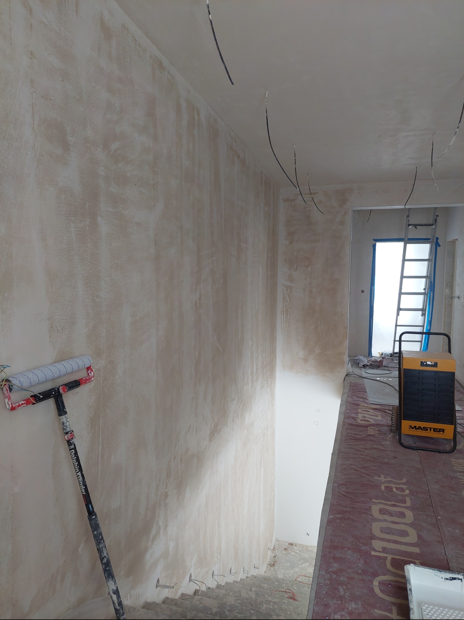 Ściana po szpachlowaniu, z widocznym wałkiem malarskim opartym o ścianę. W tle drabina i nagrzewnica Master. Widoczne kable wystające ze ściany.