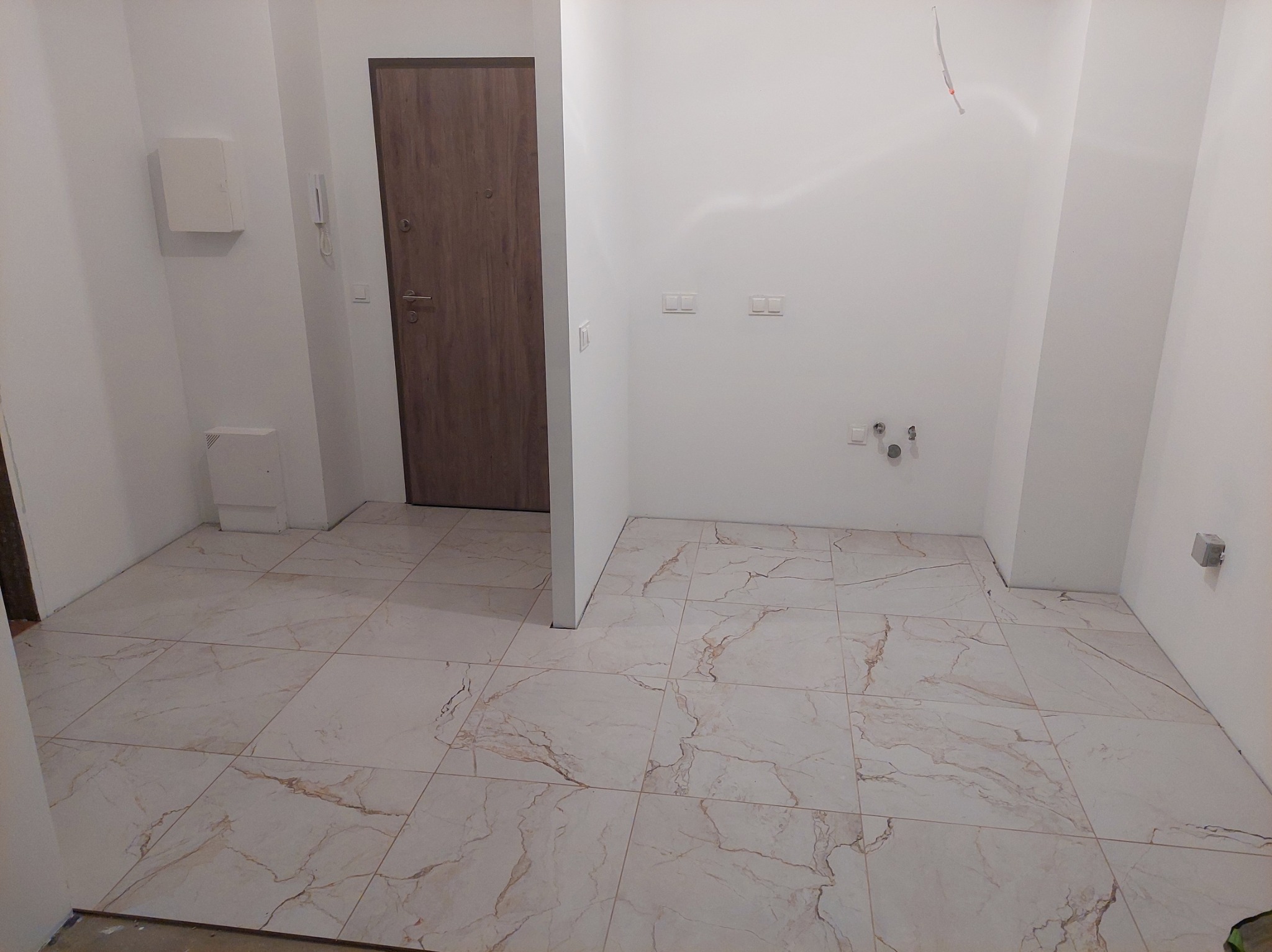 Wykończone wnętrze z beżową podłogą imitującą marmur, białe ściany z instalacjami i brązowe drzwi z metalowymi elementami w nowym mieszkaniu.
