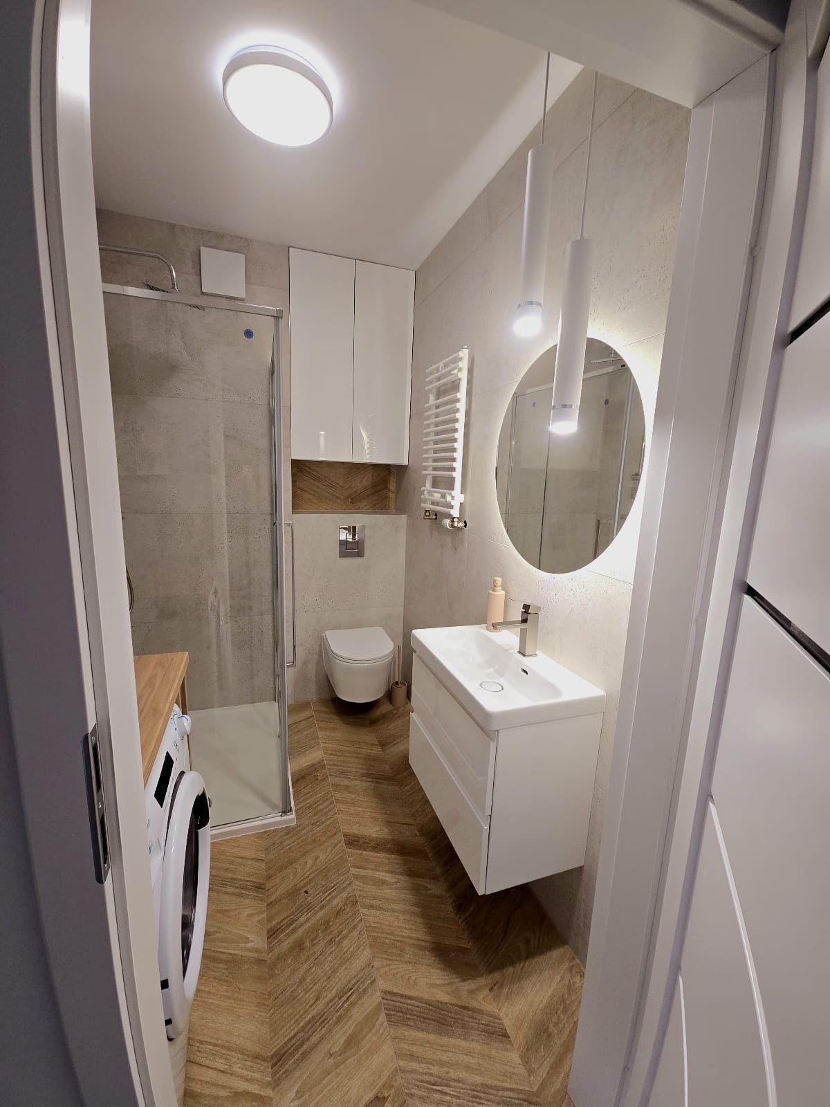 Nowoczesna łazienka z kabiną prysznicową, umywalką z szafką i drewnopodobną podłogą ułożoną w jodełkę. Oświetlenie punktowe i okrągłe lustro z podświetleniem LED.